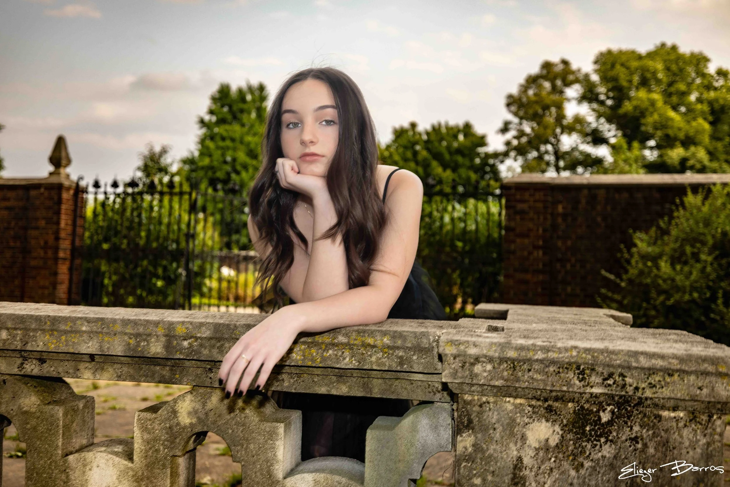 A young woman with long brown hair wearing a black dress and a ring on her finger, resting her chin on her left hand while sitting at a stone table outdoors, with a background of trees, brick wall, and a black wrought iron fence.