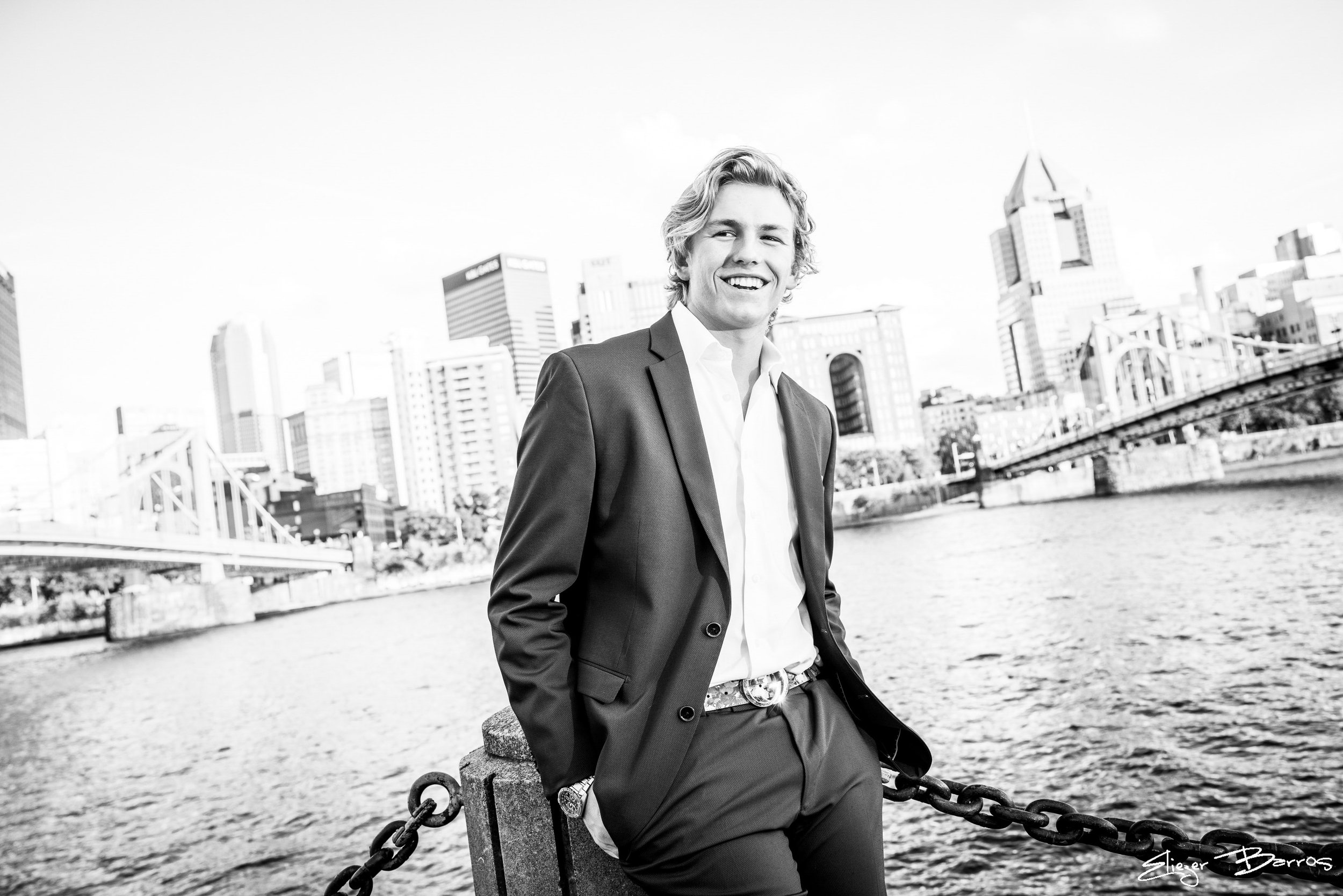 Black and white photo of a smiling young man in a suit standing outdoors overlooking a city skyline with tall buildings and a river, near a bench and chain barrier.