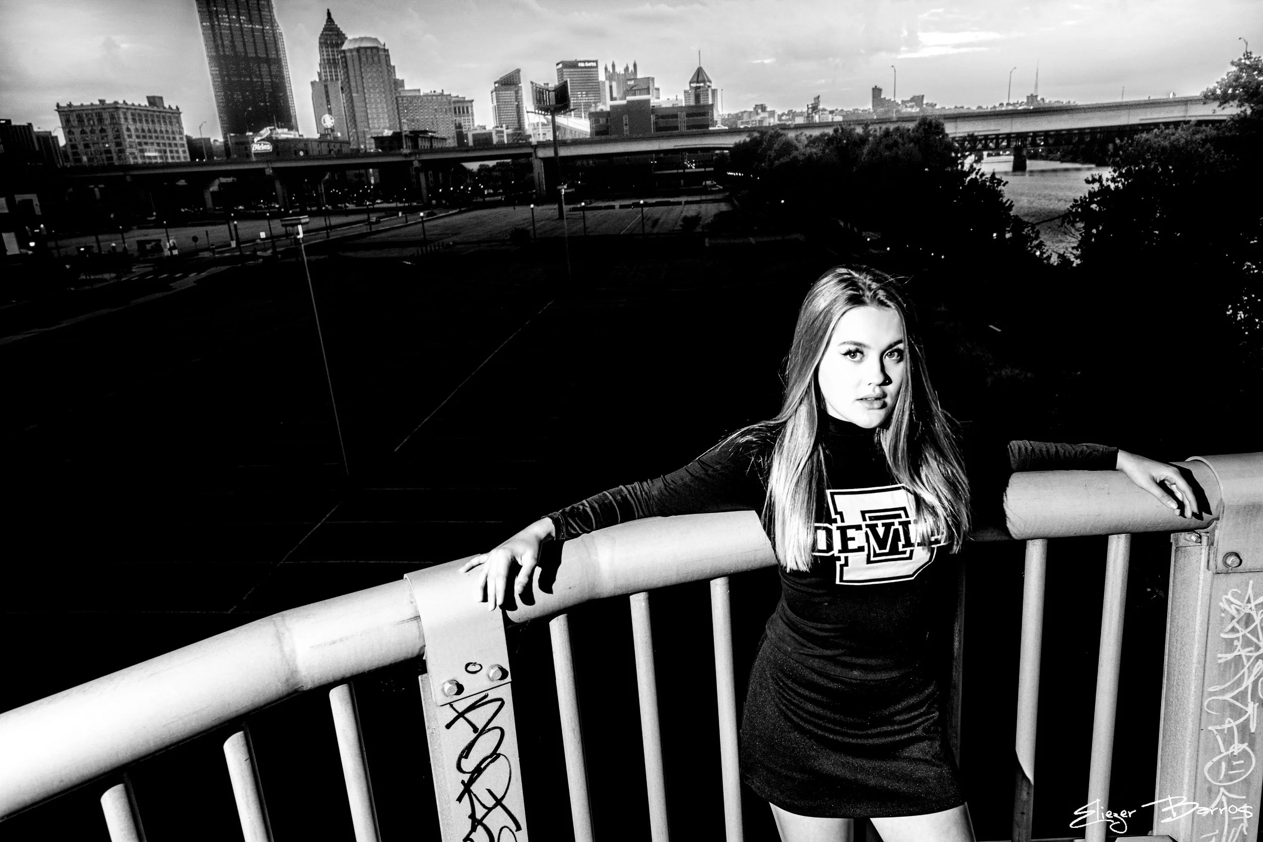 A young woman with long hair standing outdoors on a bridge railing with a city skyline in the background, dressed in a black long-sleeve top and shorts.