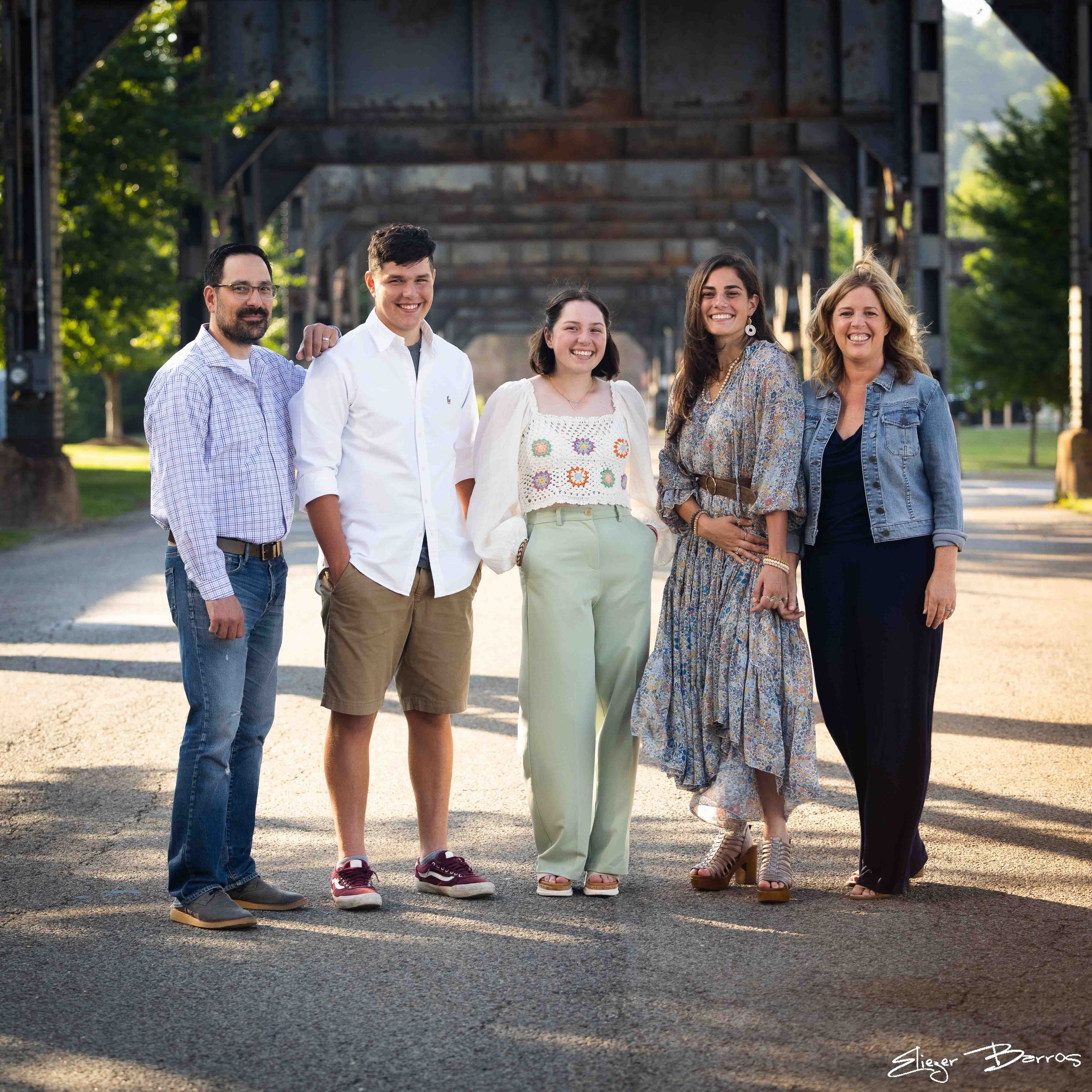 Group of five people standing outdoors under a bridge, smiling and posing for the photo. They are dressed casually, with trees and greenery in the background.