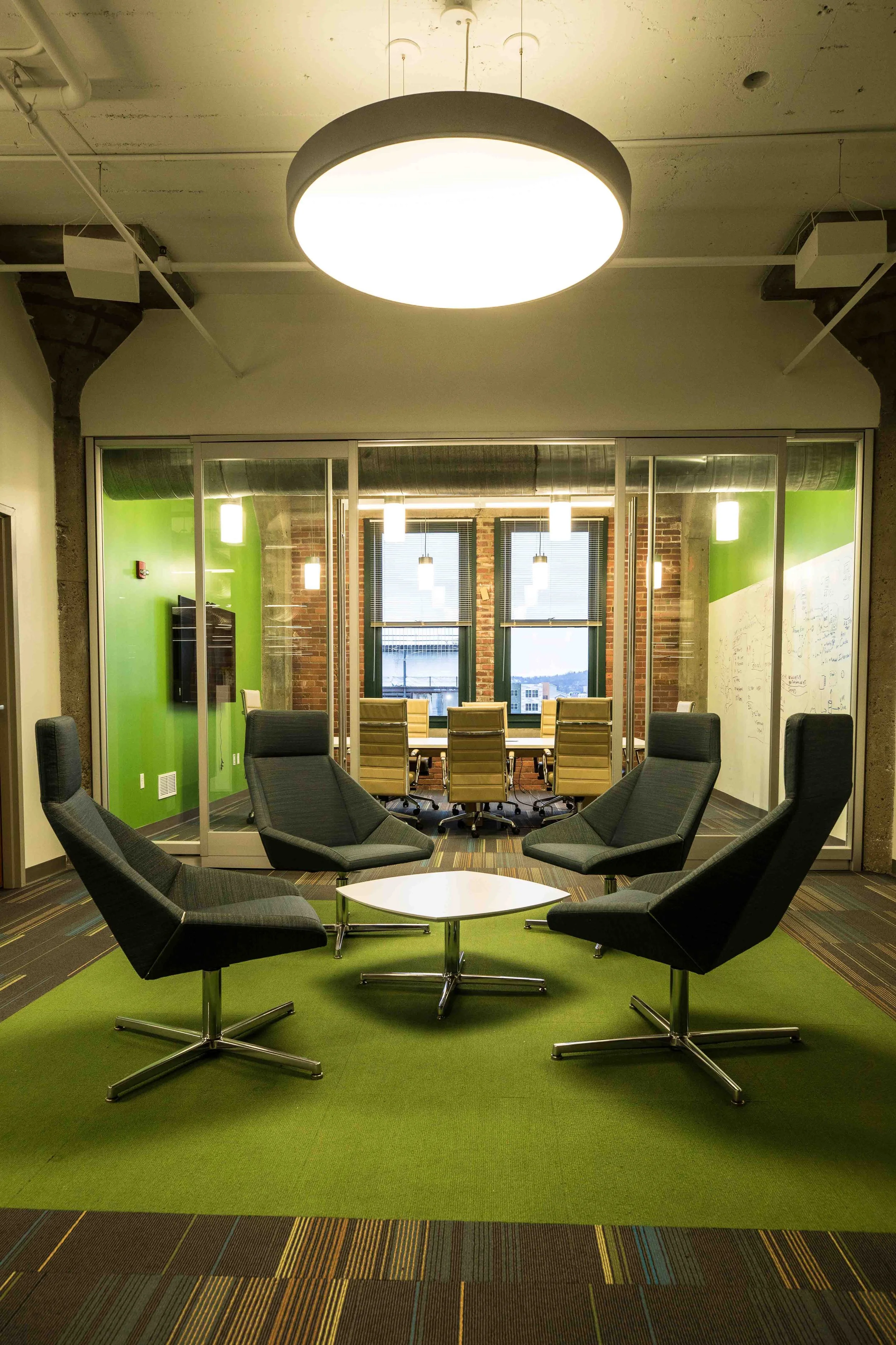 A modern office conference room with a large central round ceiling light, black swivel chairs arranged in a circle on green carpet, glass-walled room with beige chairs, and a view of city buildings through windows in the background.