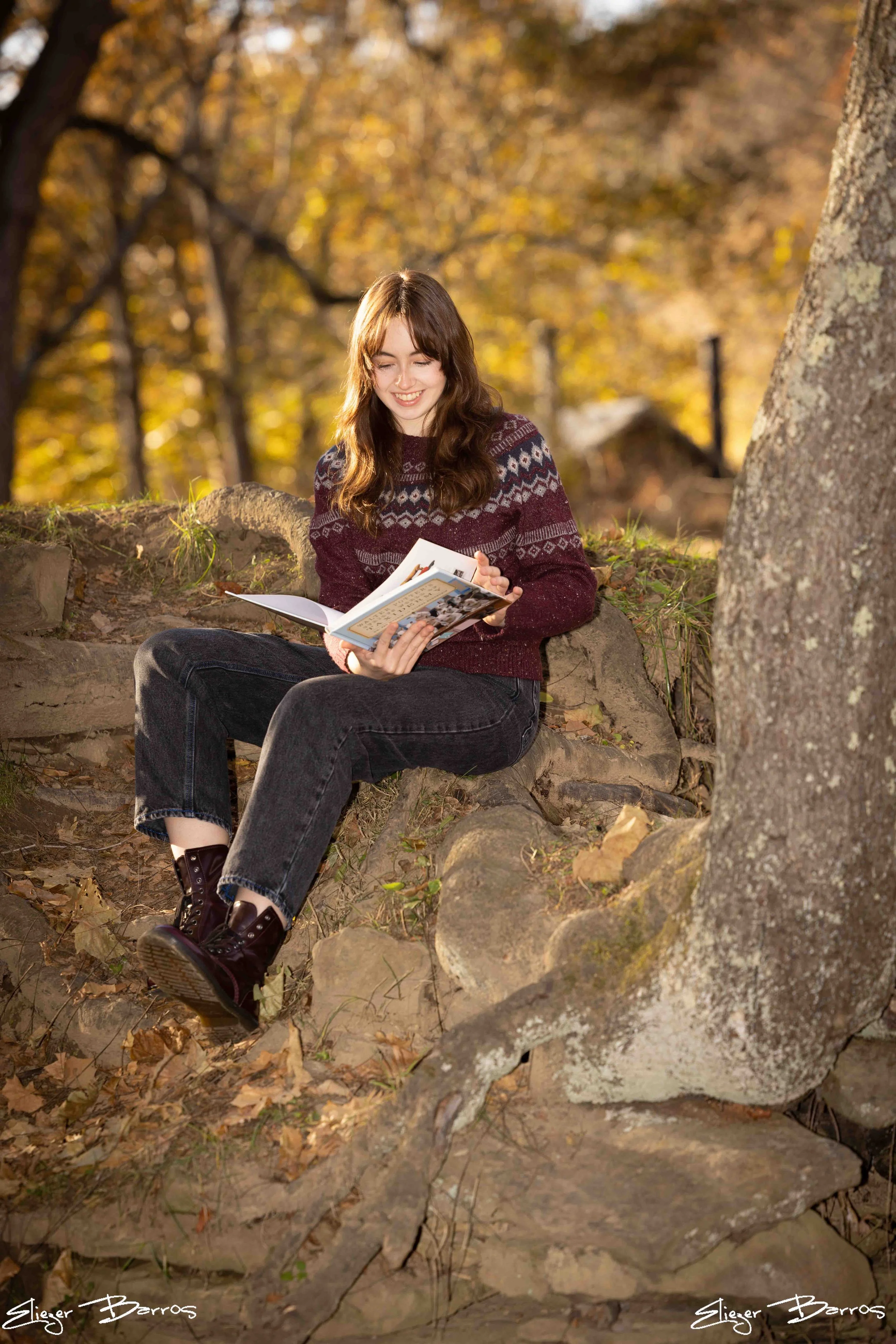 A young woman with long brown hair sitting on a rock in a forest during autumn, reading a book and smiling.