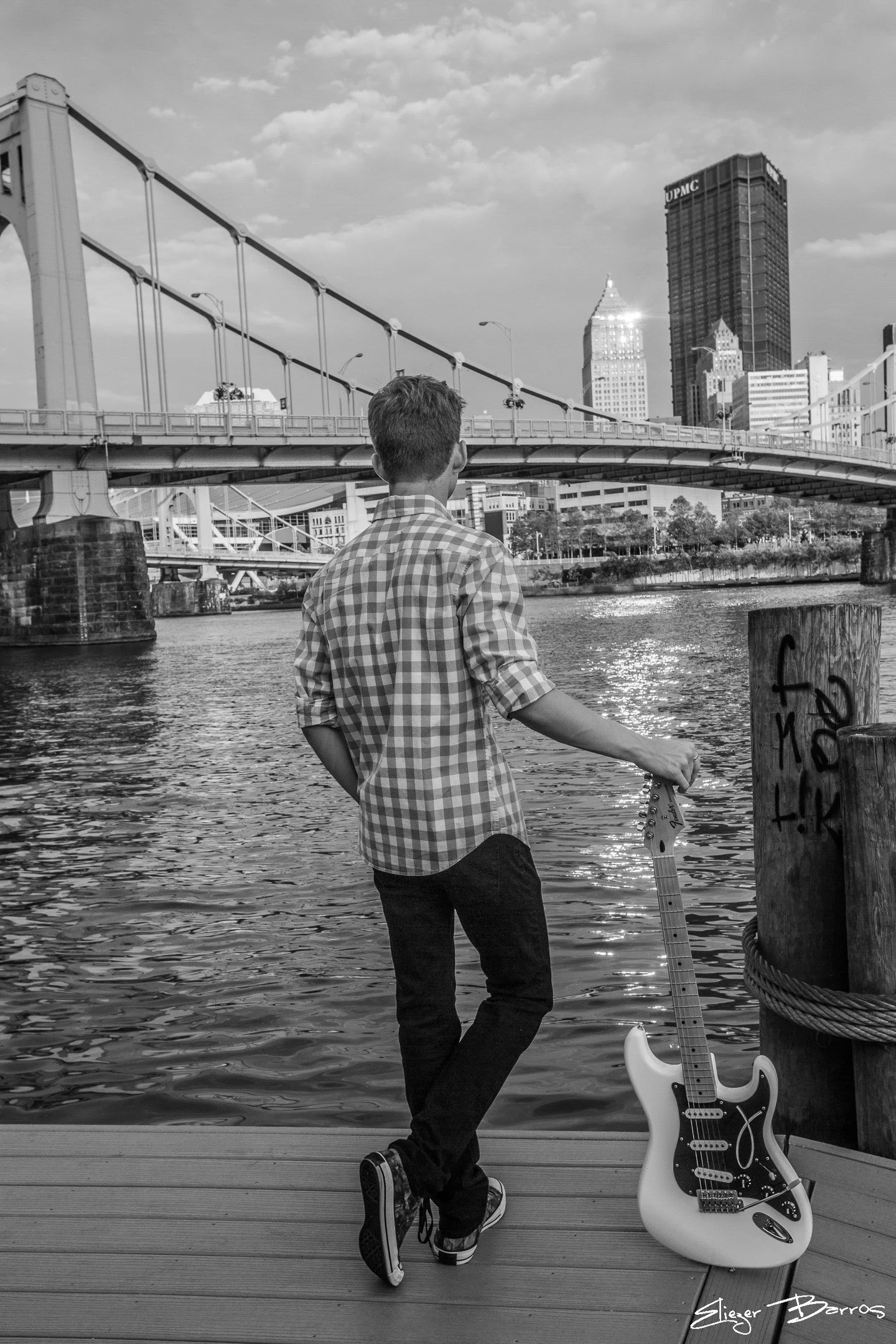 A young man with short hair, wearing a checkered shirt and dark pants, stands on a dock by the water, holding an electric guitar leaning against a post with graffiti. Behind him, there is a bridge, tall skyscrapers with one labeled 'UPMC', and a part