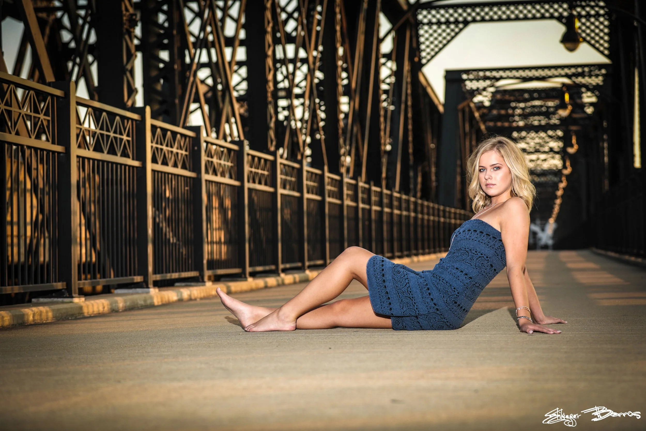A young woman with blonde hair in a blue crocheted dress sitting on a bridge at sunset.