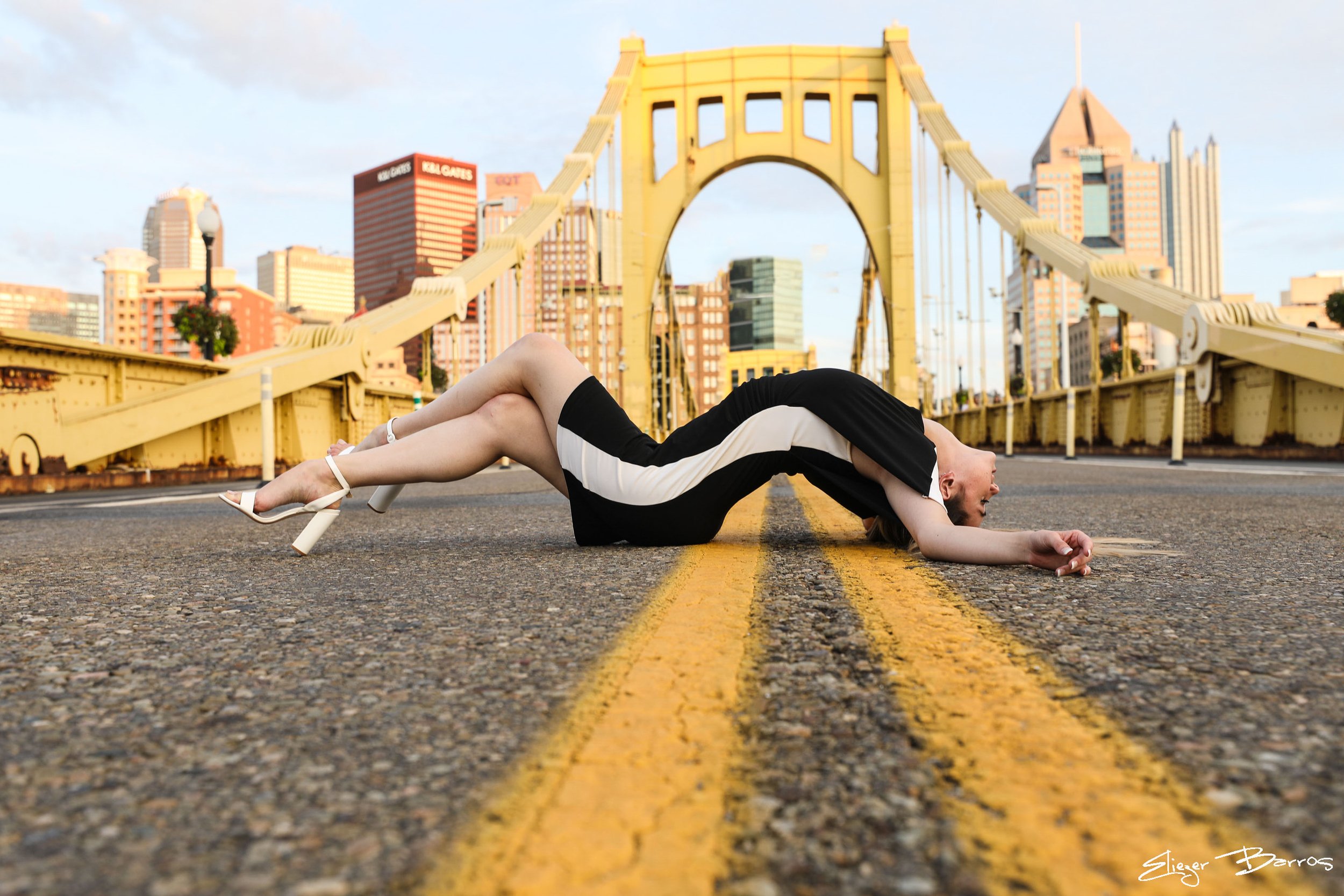 A woman lying on a city street with her upper body and head on the pavement, in front of a yellow bridge and city skyline, wearing a black and white dress and white high heels.