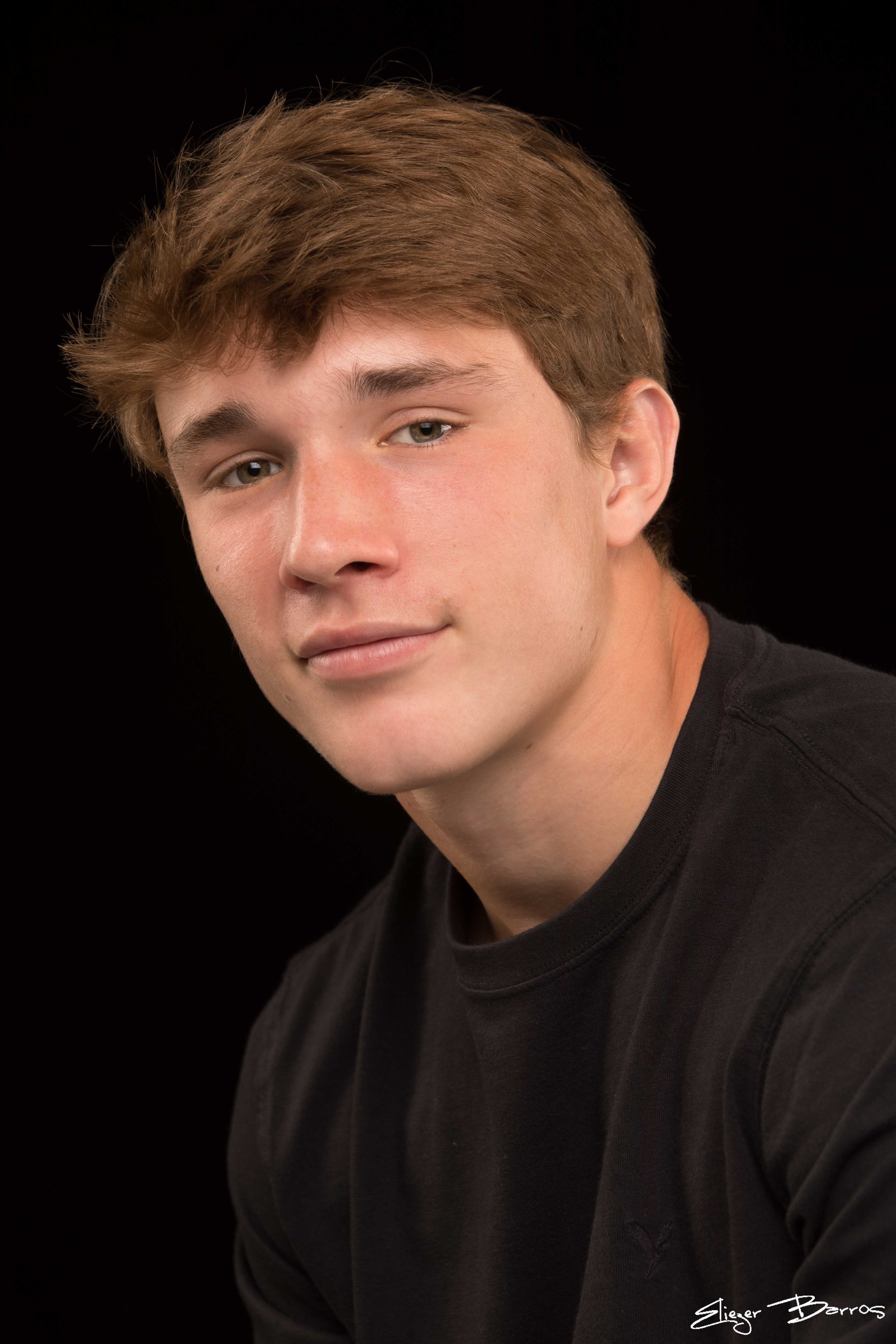 Close-up of a young man with light brown hair, blue eyes, and fair skin, wearing a black shirt against a black background.