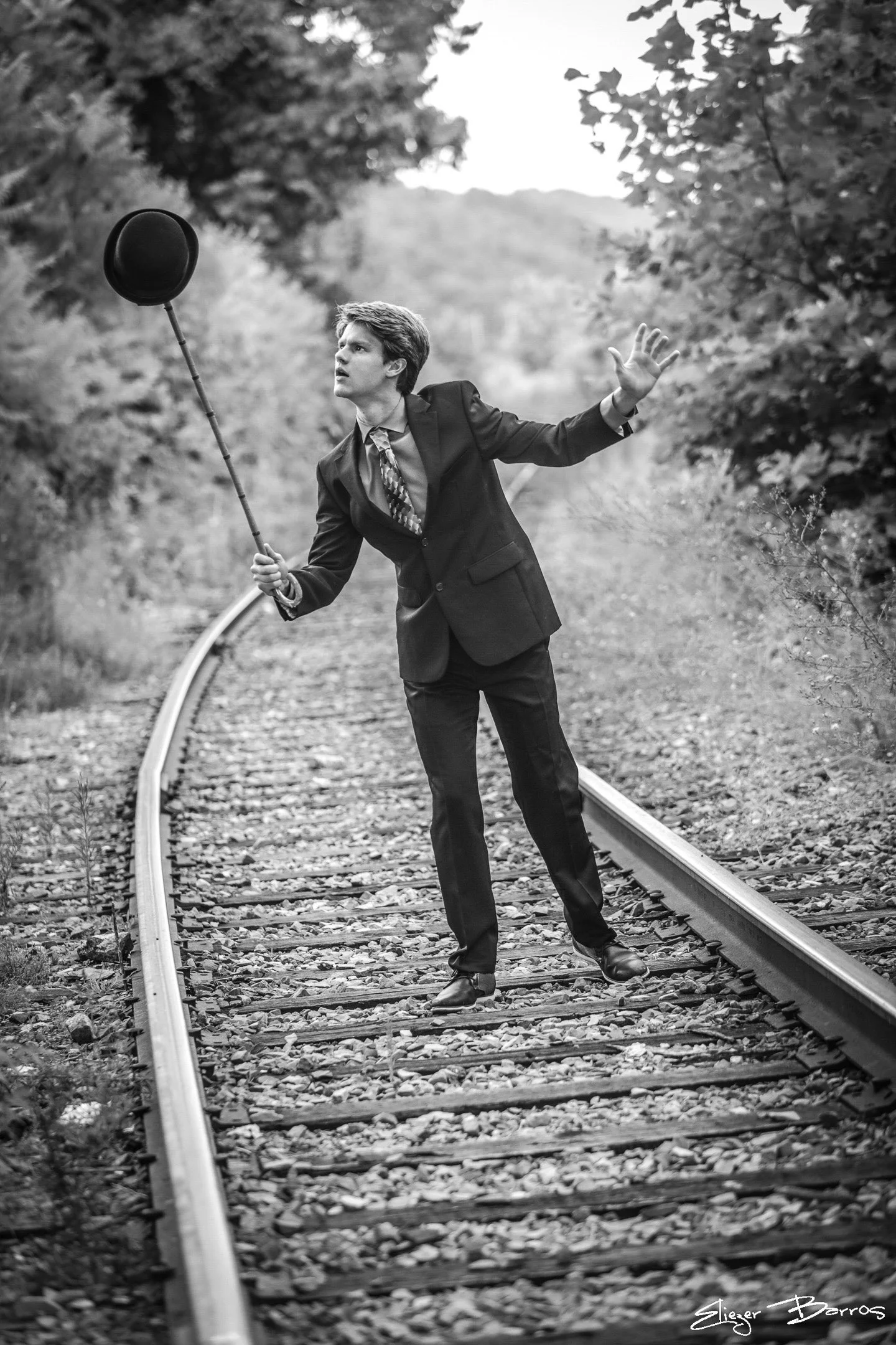 A man in a business suit holding a hat on a stick and raising his hand while walking on a railroad track in a forested area.