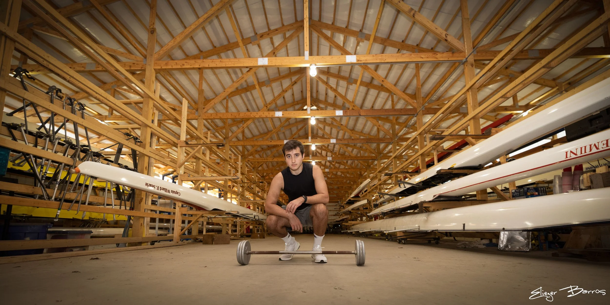 A man crouching on the floor in a wooden boat storage area with boats on racks, holding a barbell in front of him, wearing a black sleeveless shirt and gray shorts.