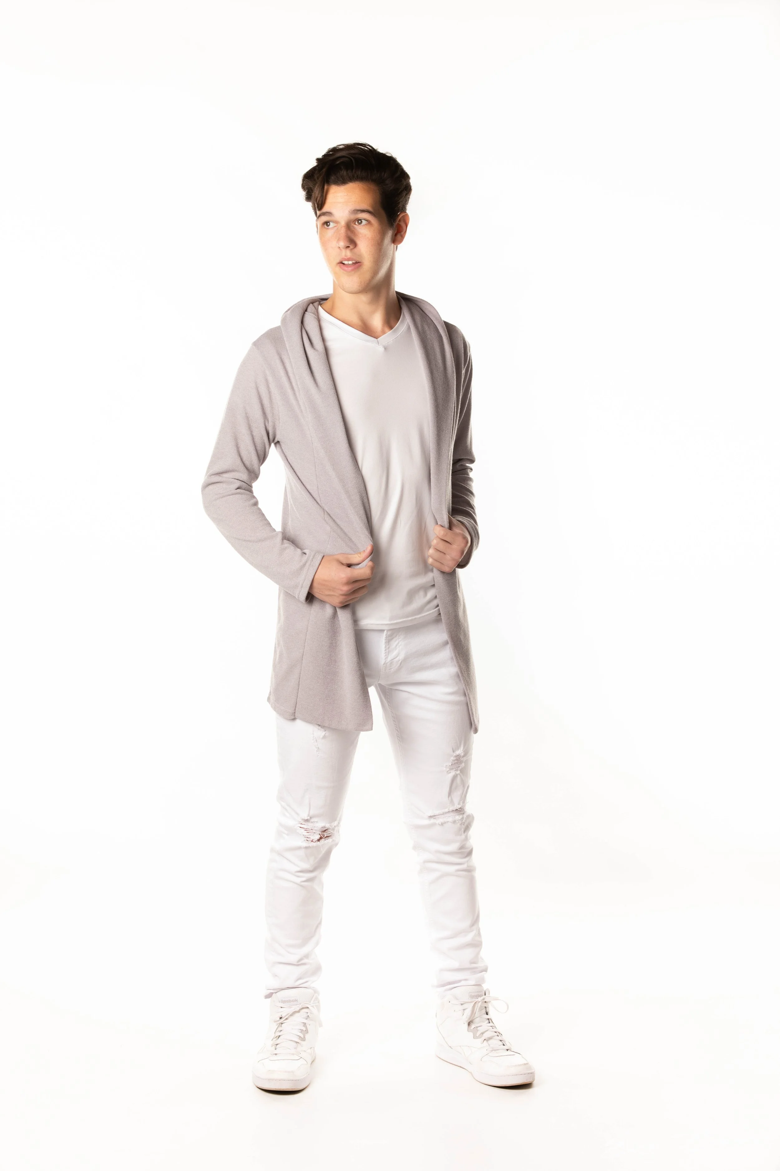 A young man with dark hair wearing a white shirt, light gray jacket draped over his shoulders, white ripped jeans, and white sneakers, standing against a white background.