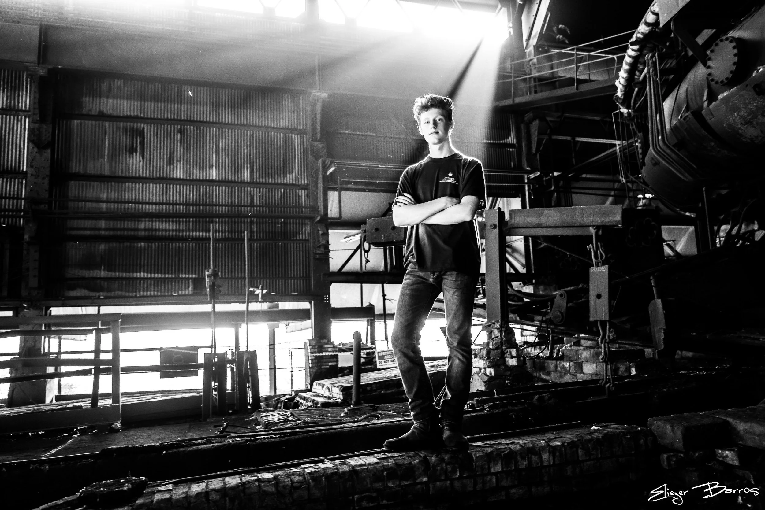 A person standing with arms crossed inside an industrial or construction setting, with metal structures and equipment around. Sunlight streams through the space, creating a bright spot at the top left. The photo is in black and white.