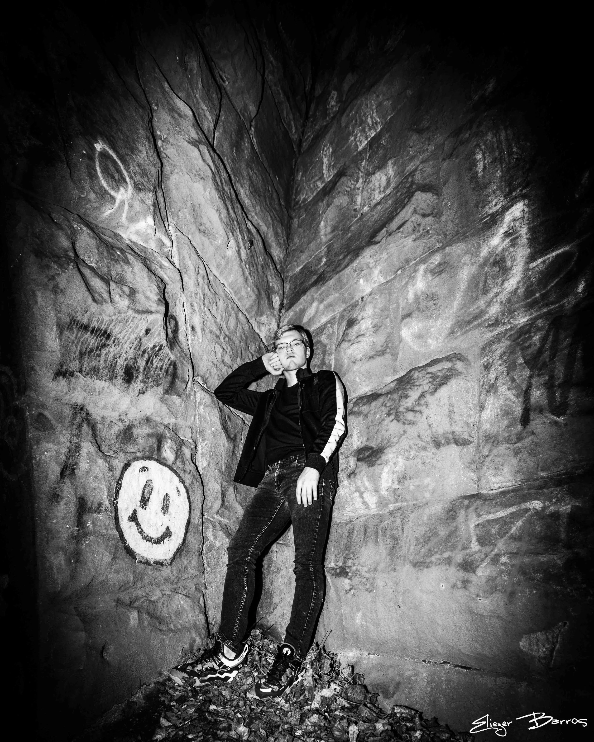 A young man wearing glasses and a jacket stands in front of a large rock formation with graffiti and smiley face drawings, with dried leaves on the ground, captured in black and white.