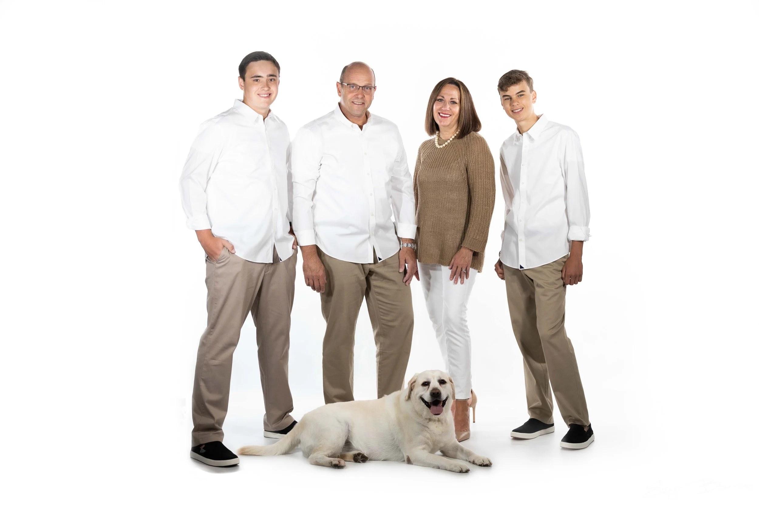 A family of four standing with a Labrador Retriever dog in front of a plain white background. The family includes two young men, a woman, and an older man, all dressed in casual white or tan clothing.
