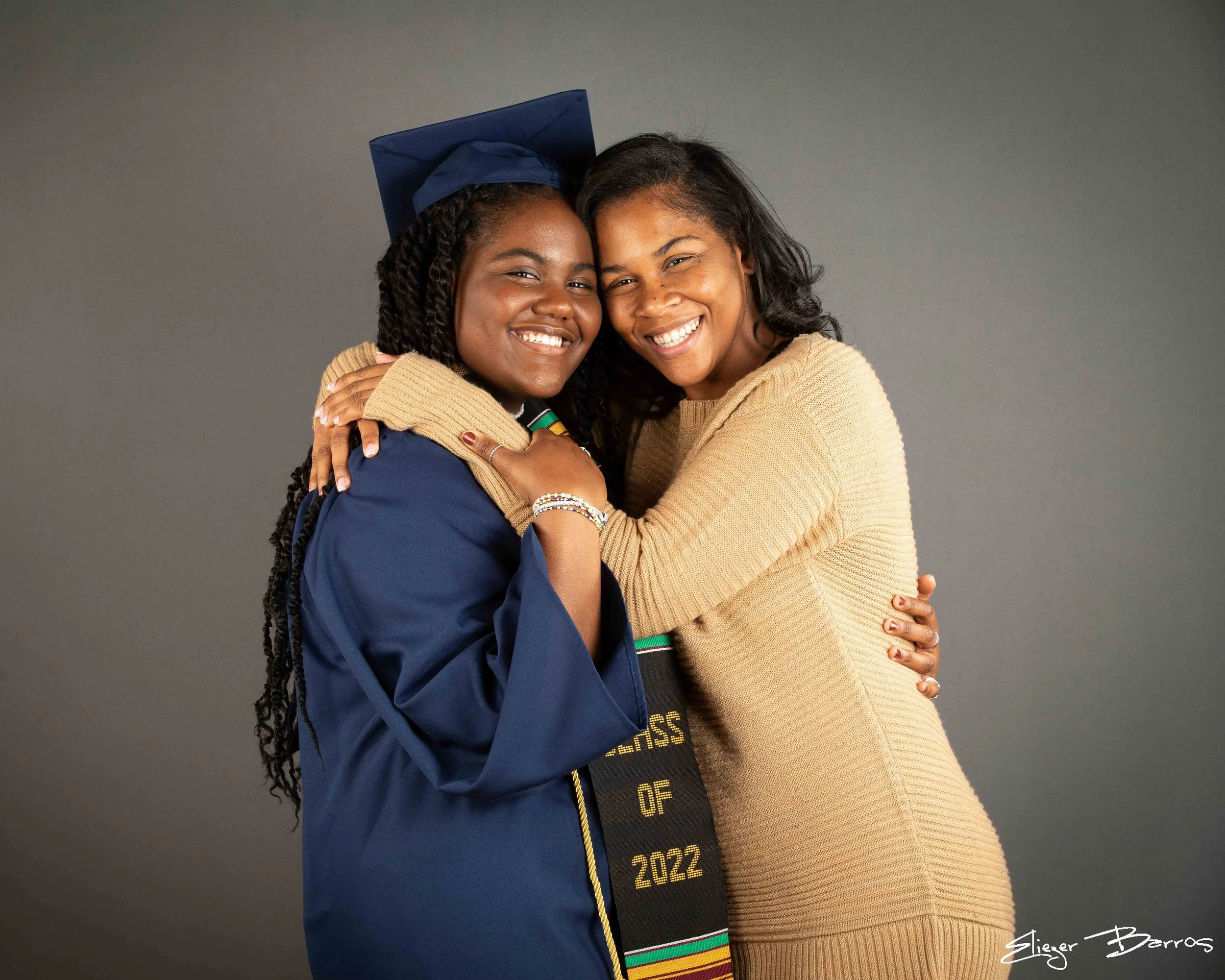 Two smiling women hugging each other. One is wearing a navy blue graduation cap and gown, the other is in a beige sweater. The background is gray. The woman in graduation attire has long braided hair and is wearing jewelry, the other has shoulder-len