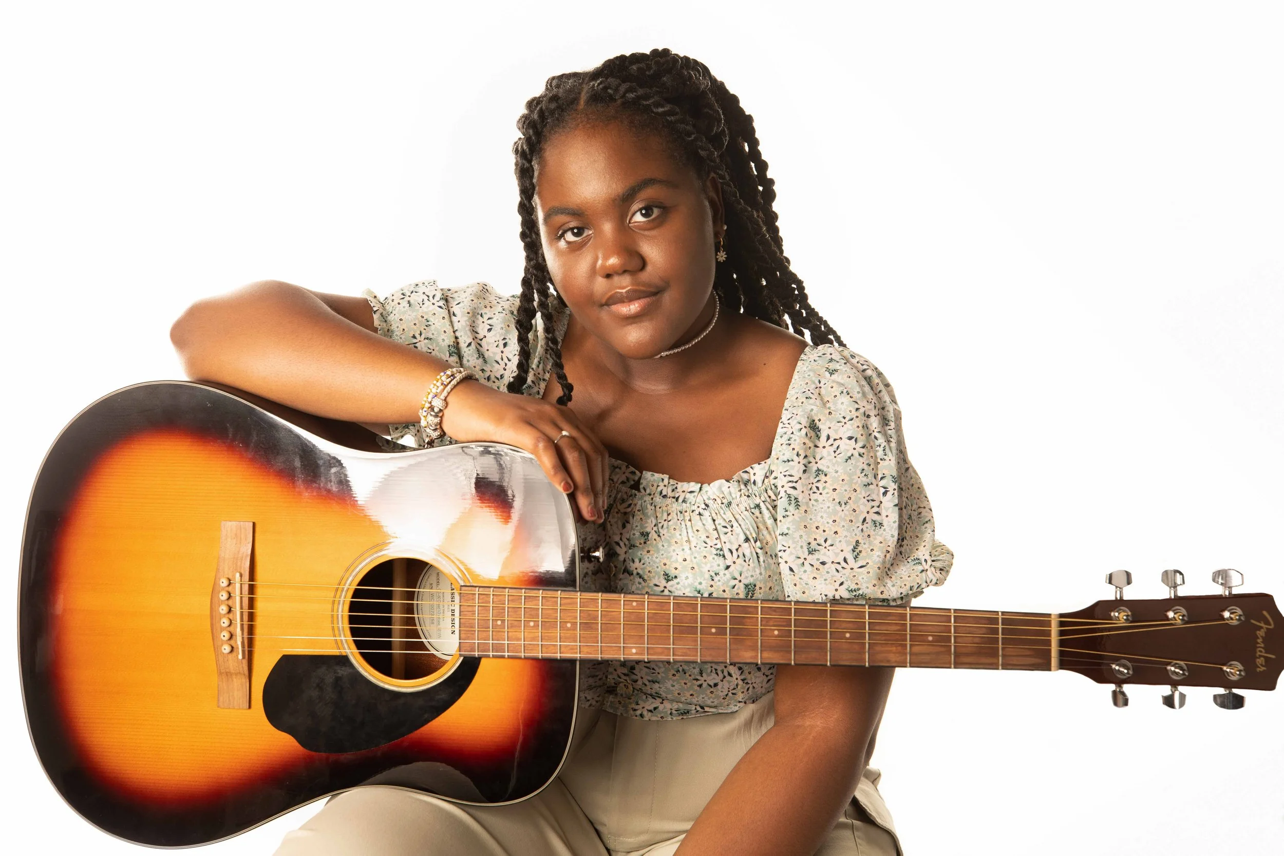 A young woman with braided hair and earrings, wearing a floral patterned top, sitting with a sunburst acoustic guitar on her lap, against a white background.