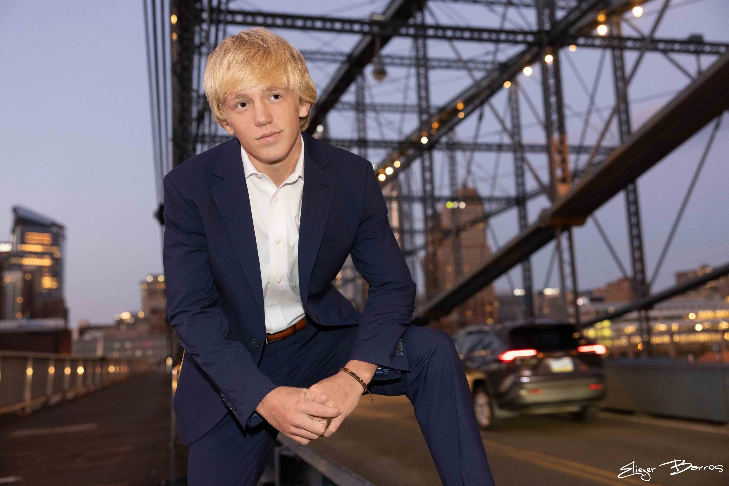 A young man in a dark blue suit sits on a railings on a rooftop with city buildings and a bridge in the background during dusk. He has blonde hair, a white shirt, and a serious expression.