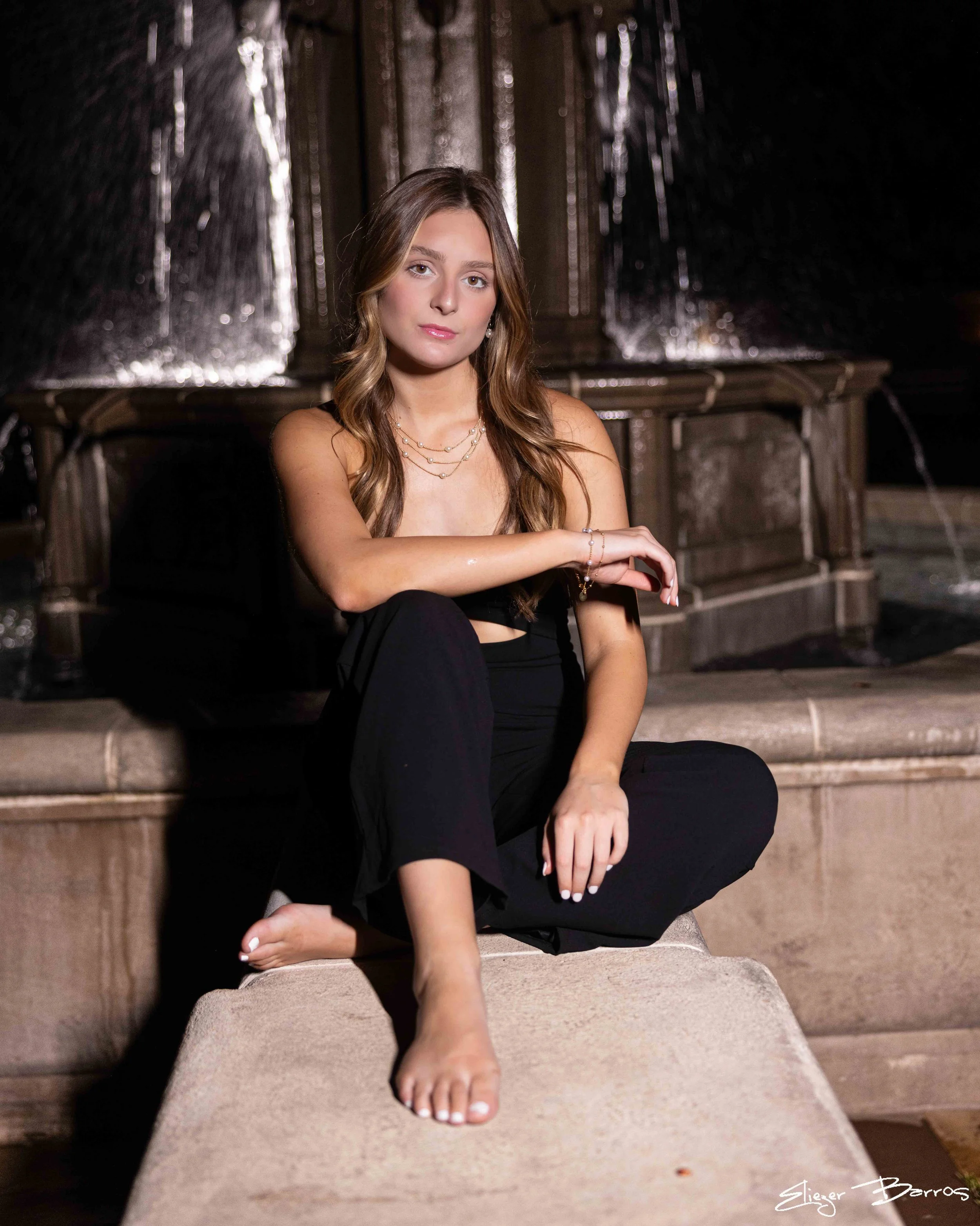 A young woman with long wavy hair, sitting on stone steps in a dimly lit setting with dark wooden background. She is dressed in black and accessorized with layered necklaces, earrings, and a bracelet, with her legs crossed and one arm resting on her 