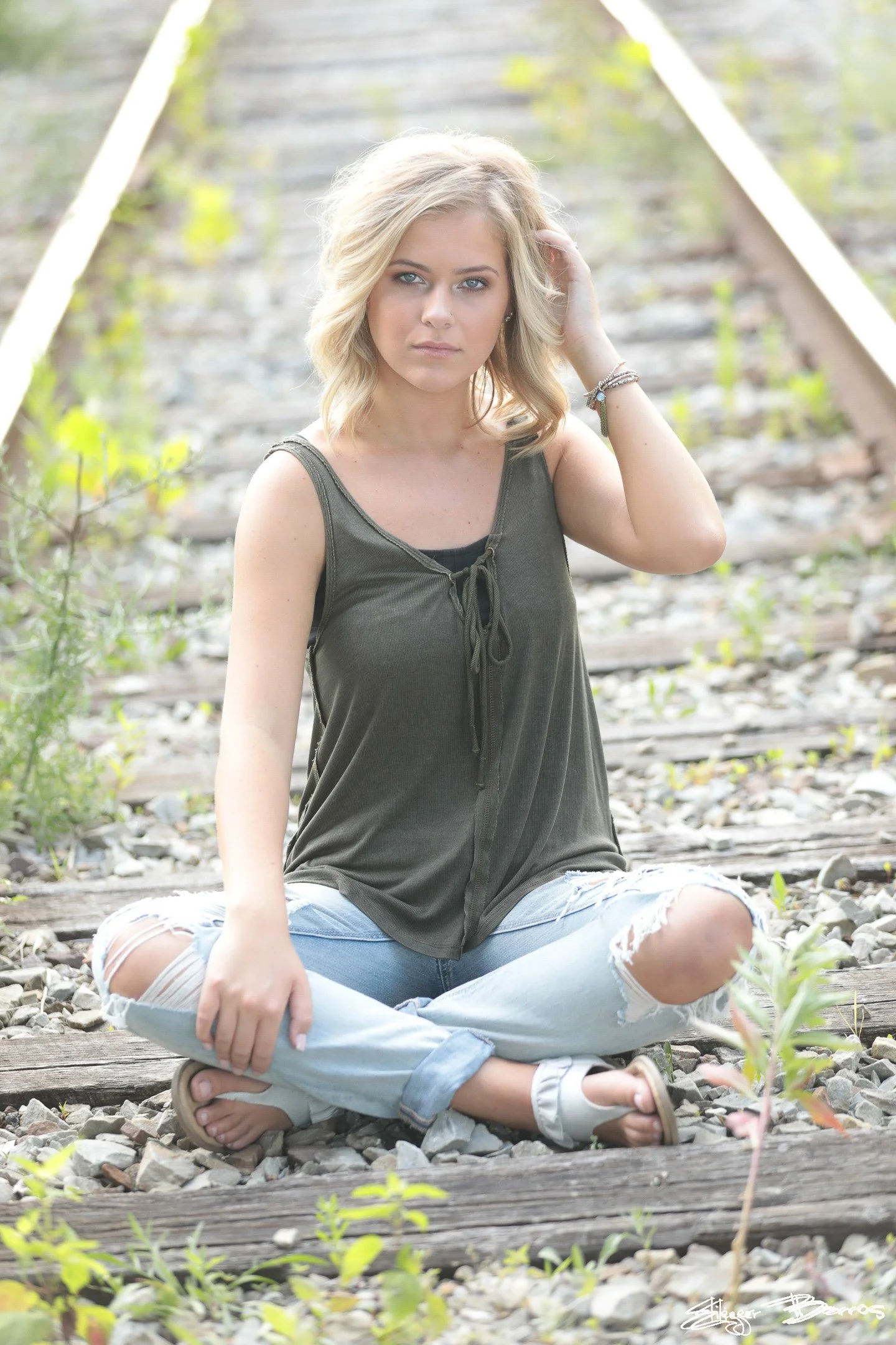 A young woman with blonde hair sitting cross-legged on a railroad track, wearing a dark sleeveless top and ripped jeans, with a neutral expression.