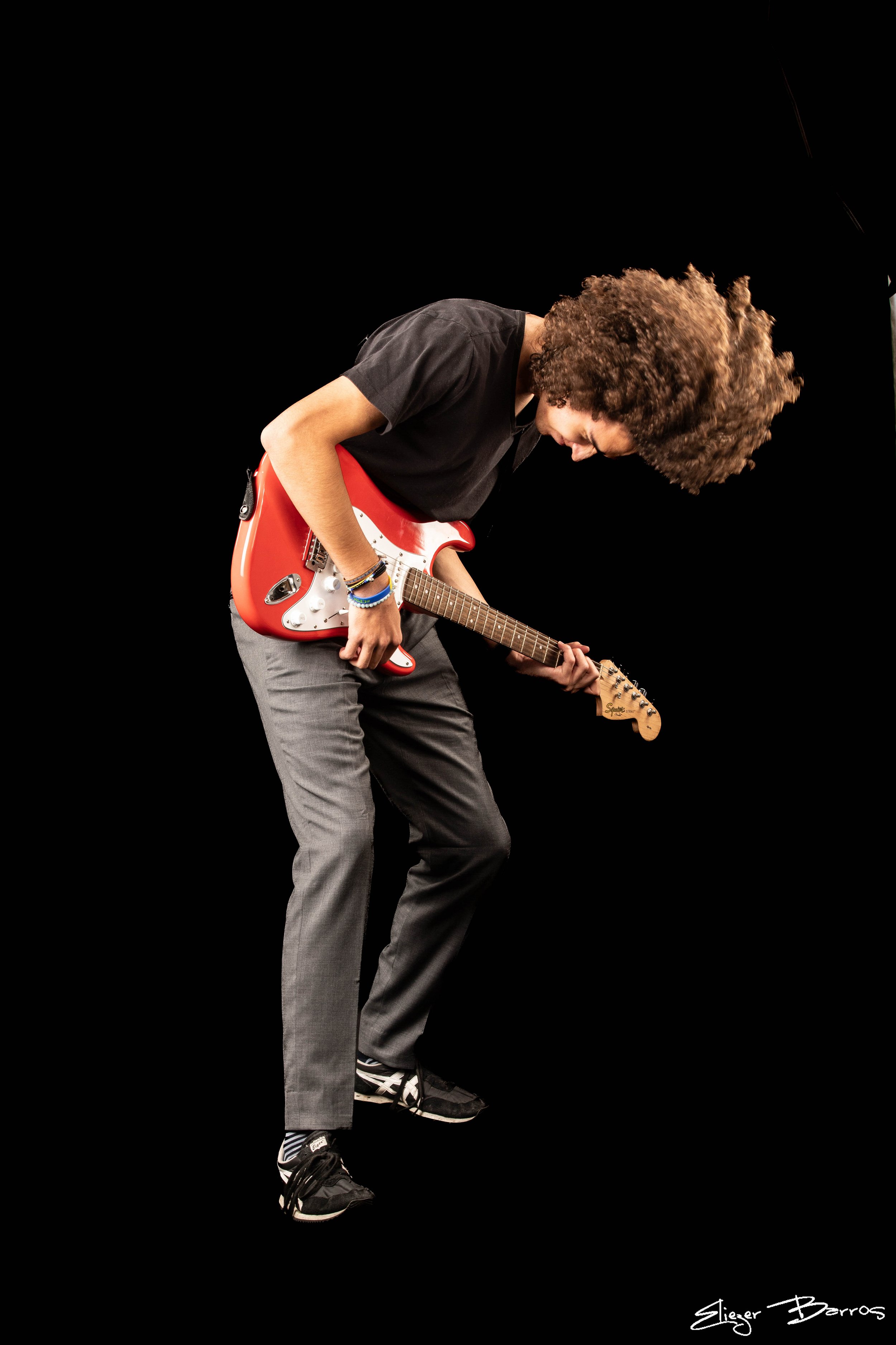 Young man with curly hair playing an electric guitar on stage against a black background, wearing a black t-shirt, gray pants, and sneakers.