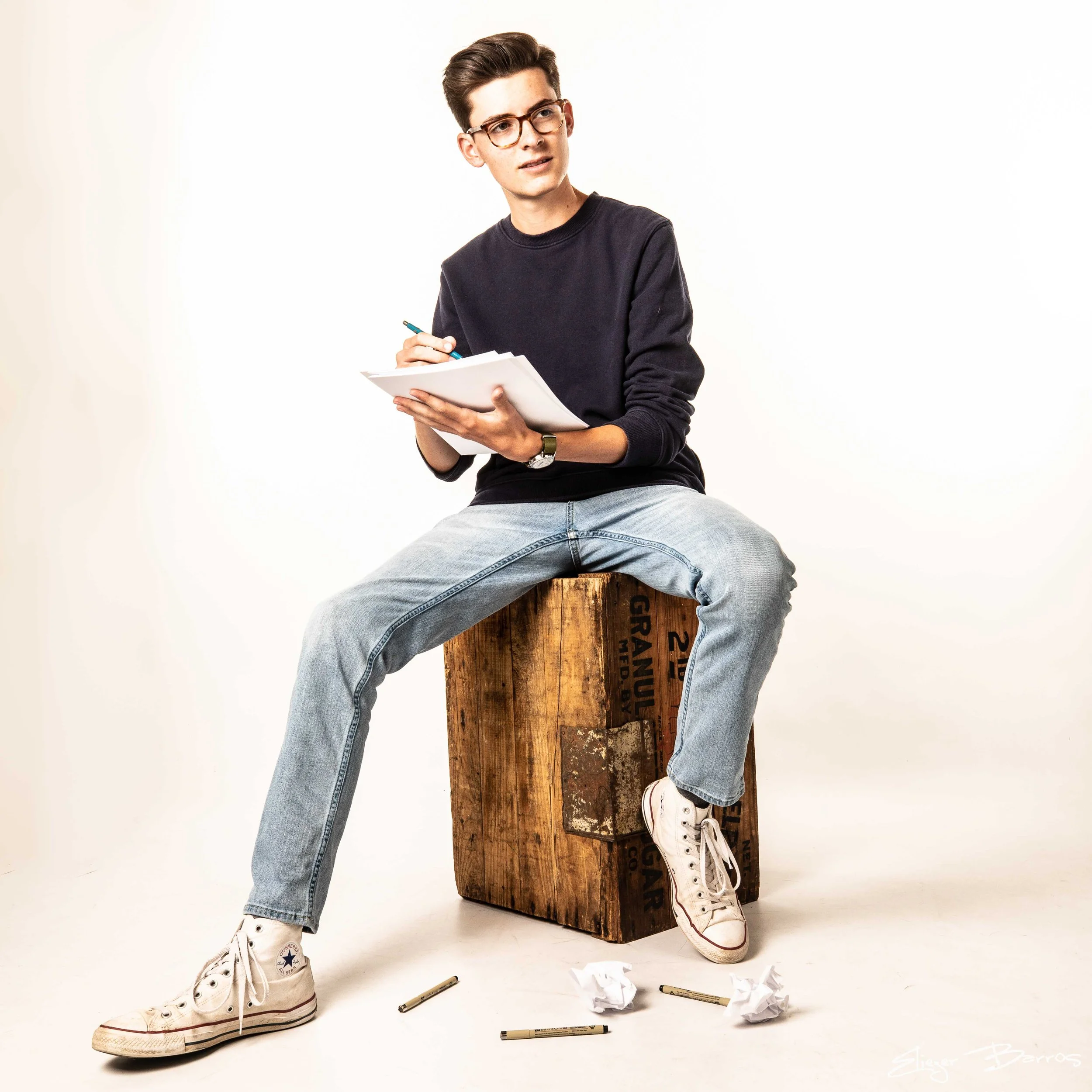 A young man sitting on a wooden crate, holding a notebook and pen, with messy crumpled papers and markers scattered on the floor, wearing glasses, a black sweater, and white sneakers.