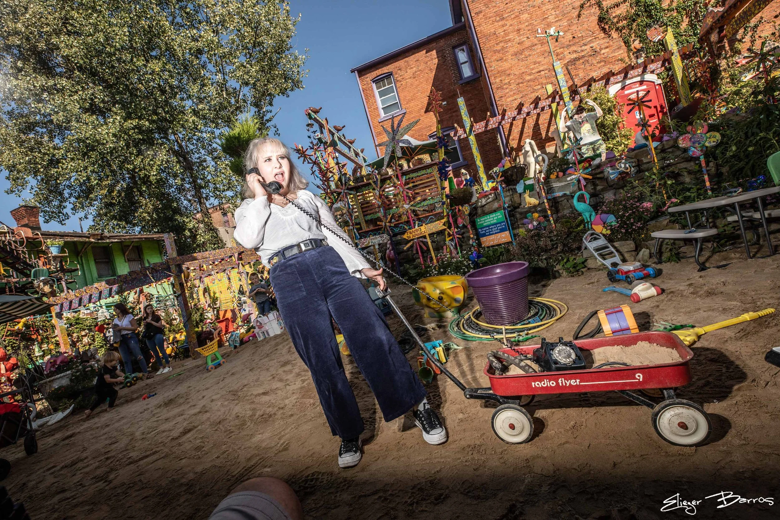 Senior Pictures Pittsburgh at Randyland Mexican War Streets. Young woman in white top and jeans pulling a red Radio Flyer wagon through a whimsical folk art wonderland for fun and artistic senior pictures.