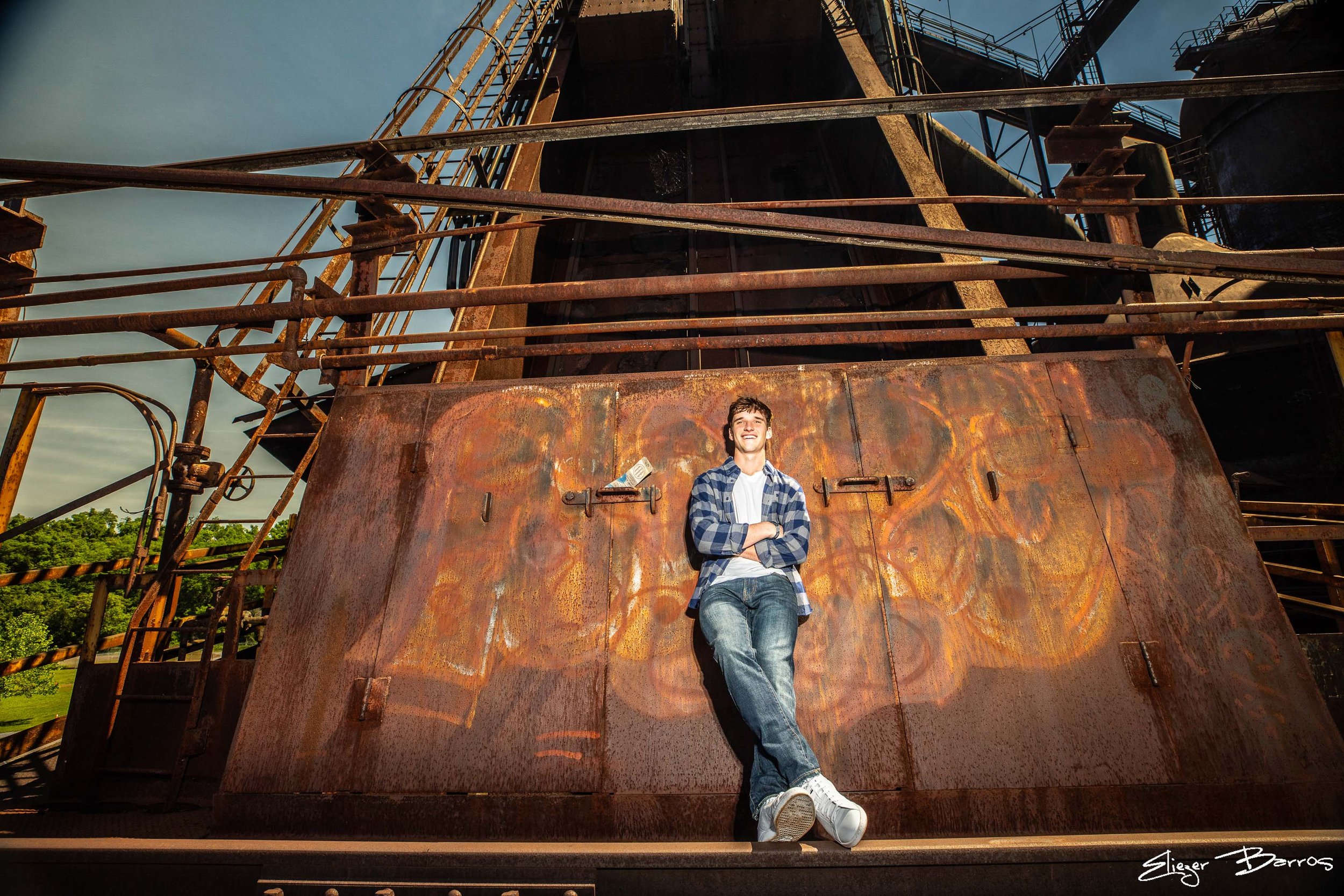 A young man in a plaid shirt and jeans leaning against a rusted metal structure with stairs and railings, smiling and crossing his arms, outdoors with trees and sky in the background.