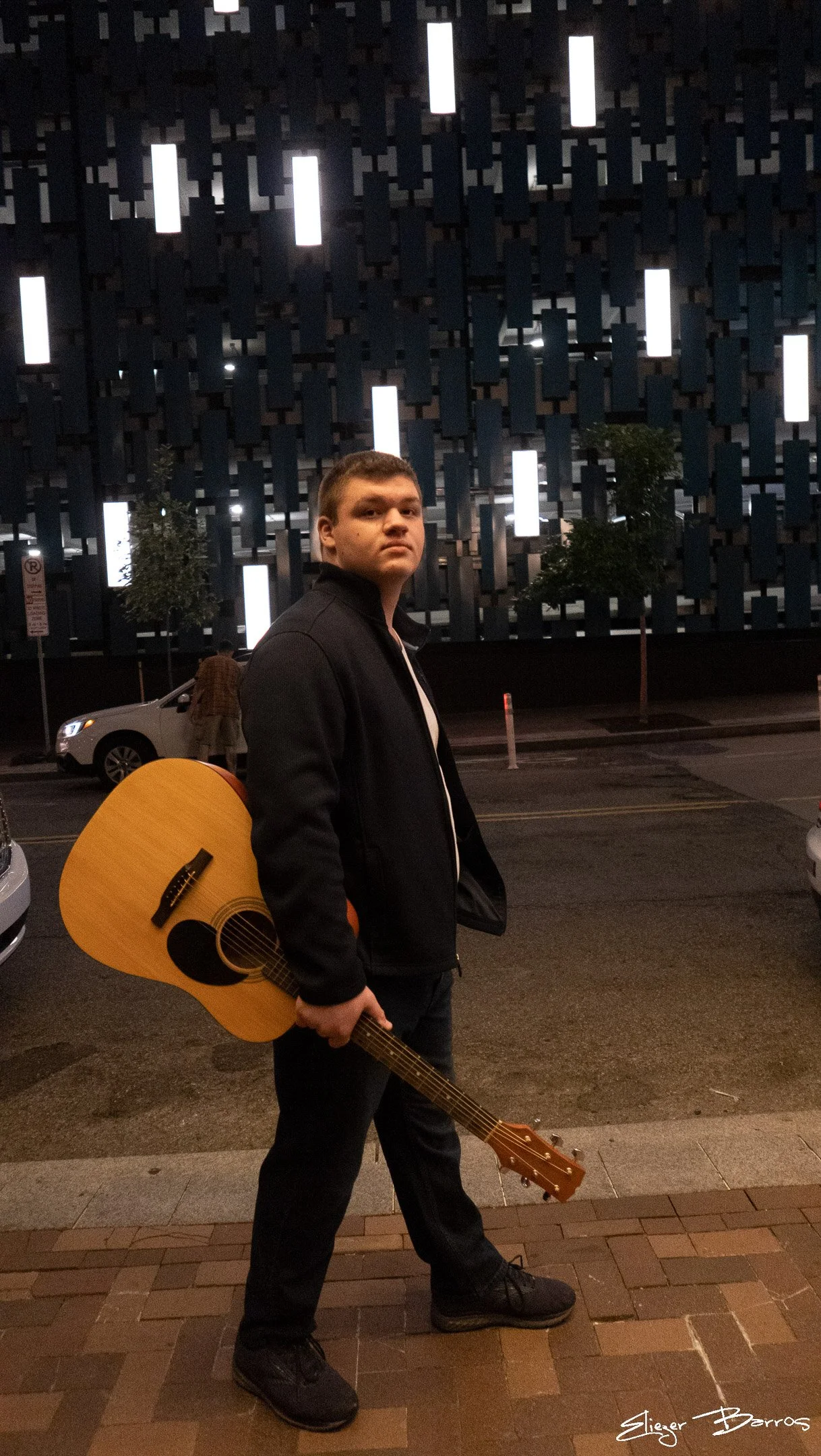 A young man stands outdoors at night holding an acoustic guitar. He is dressed in a black jacket, black pants, and black shoes. The background features a modern building with illuminated vertical slats and a couple of small trees.