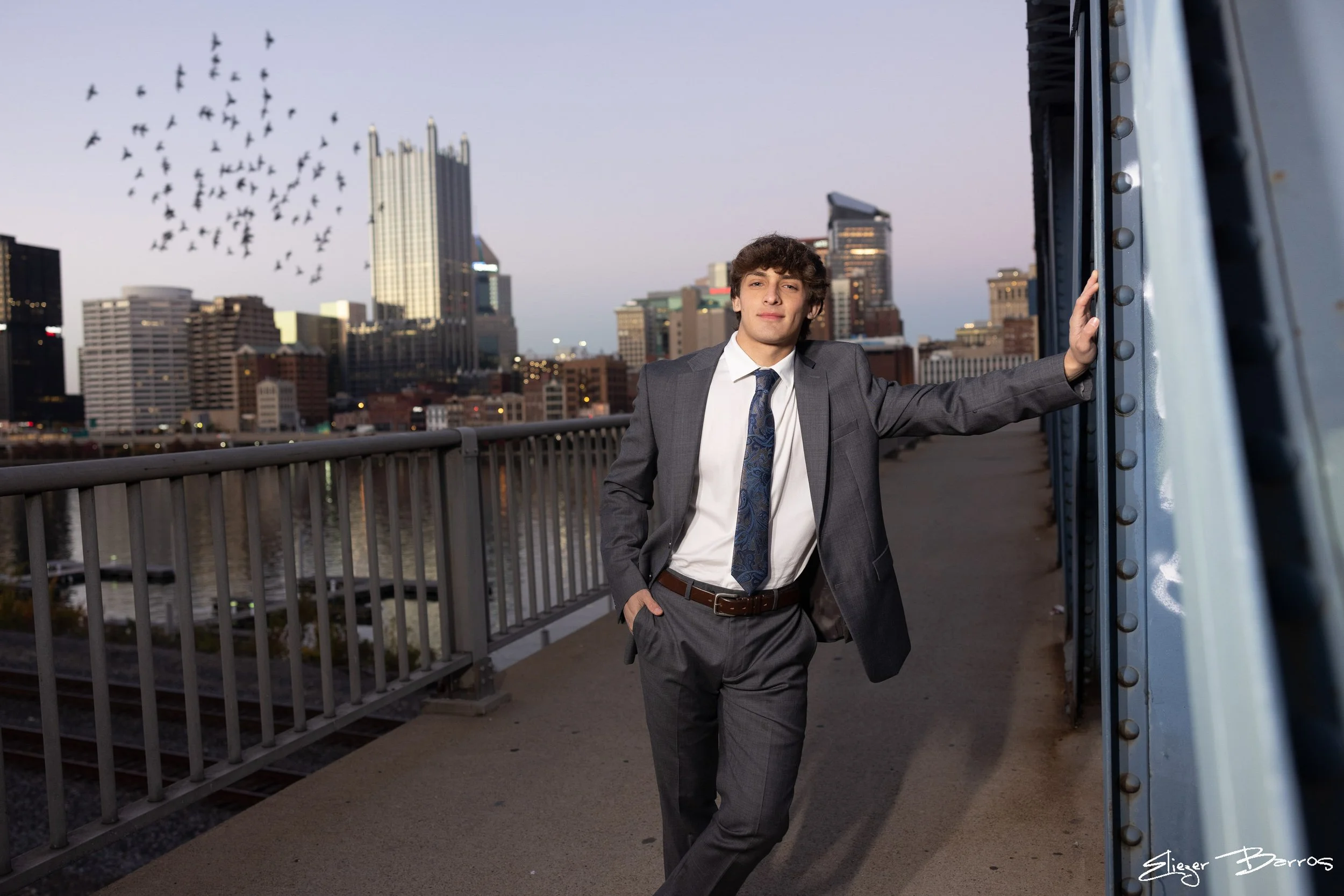A young man wearing a gray suit, white shirt, and patterned tie leaning against a metal structure on a bridge, with a city skyline in the background and a flock of birds flying in the sky.
