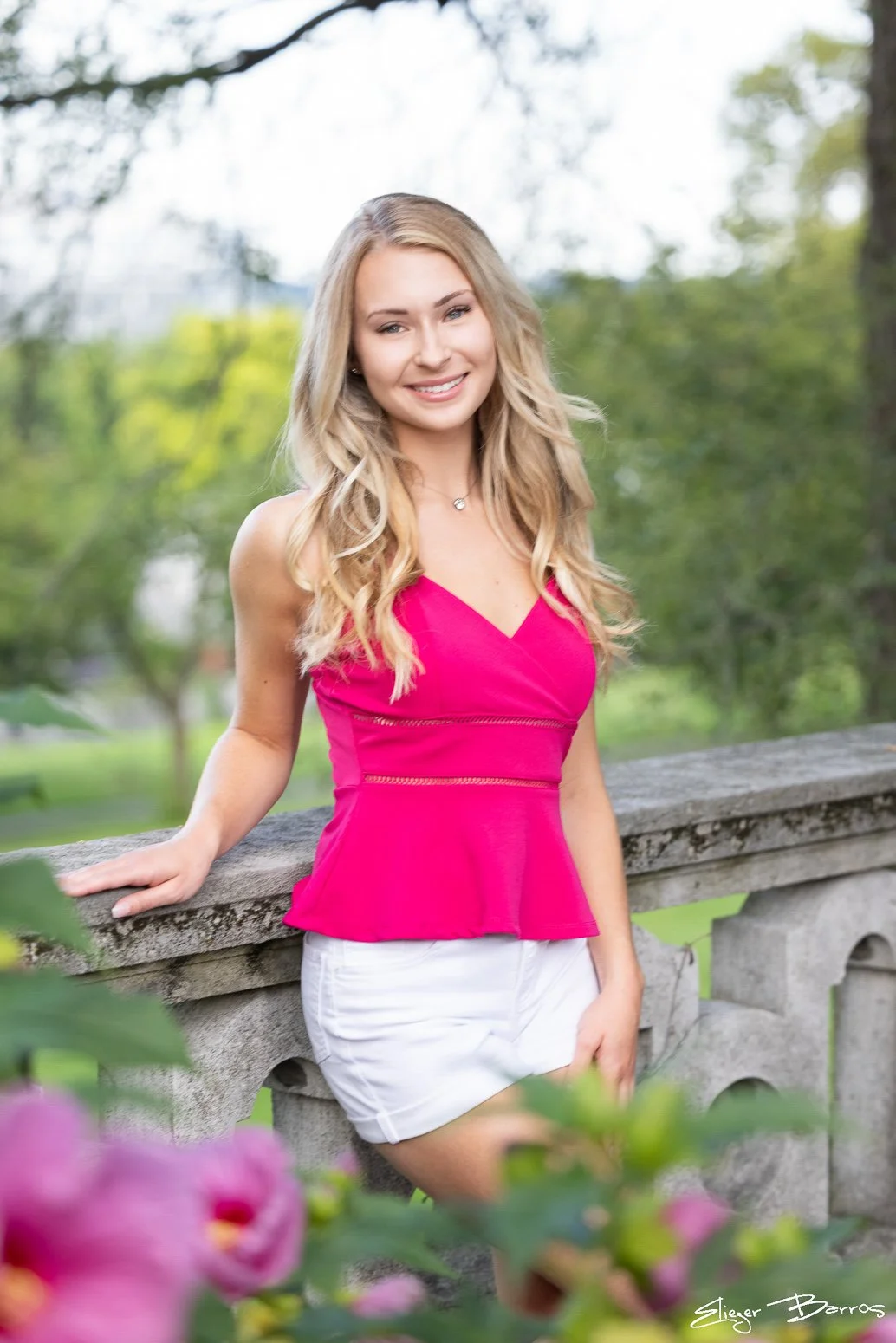 A young woman with long blonde hair wearing a bright pink sleeveless top and white shorts, standing outdoors by a stone railing with green trees and blurred background.