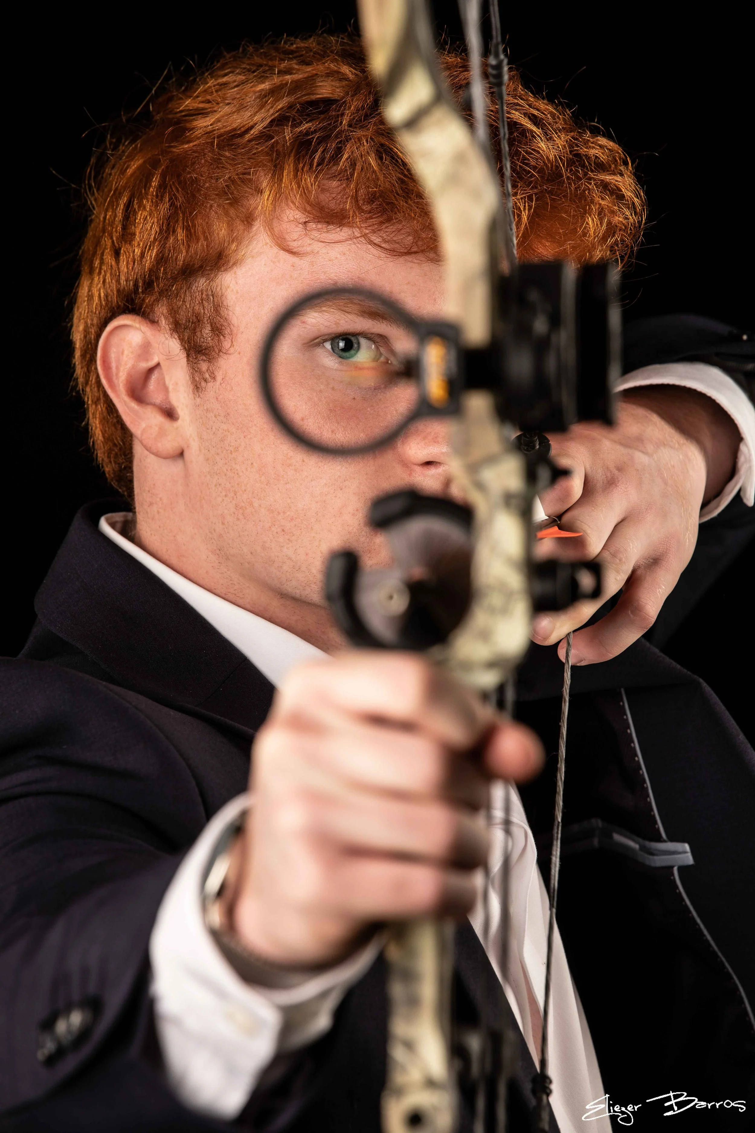A man with red hair aiming a hunting bow, wearing a dark suit with a white shirt, against a black background.