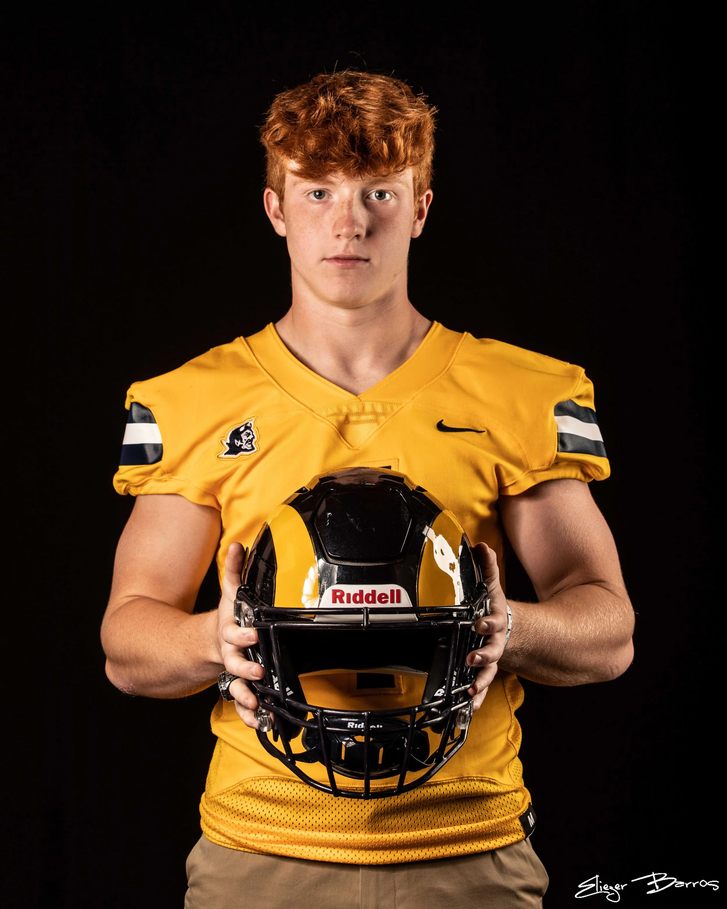 A young man with red hair wearing a yellow sports jersey and holding a black football helmet, standing against a black background.
