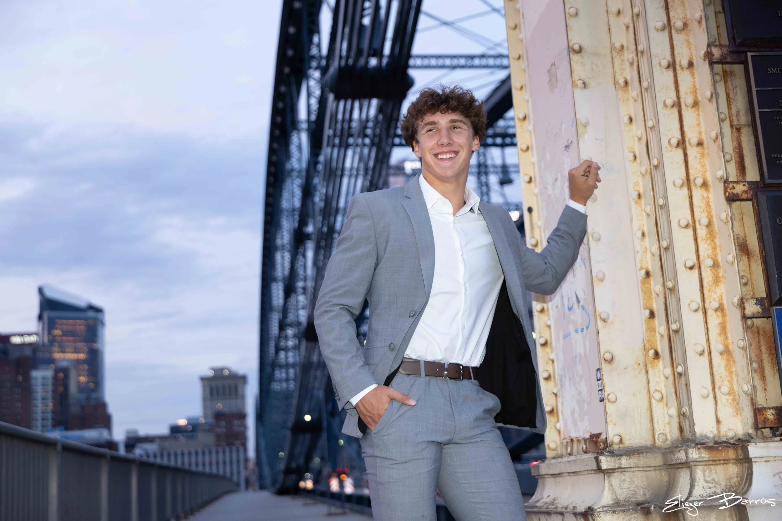 A young man in a gray suit smiling and leaning against a rusted, yellow-painted building structure on a city rooftop with buildings and cloudy sky in the background.