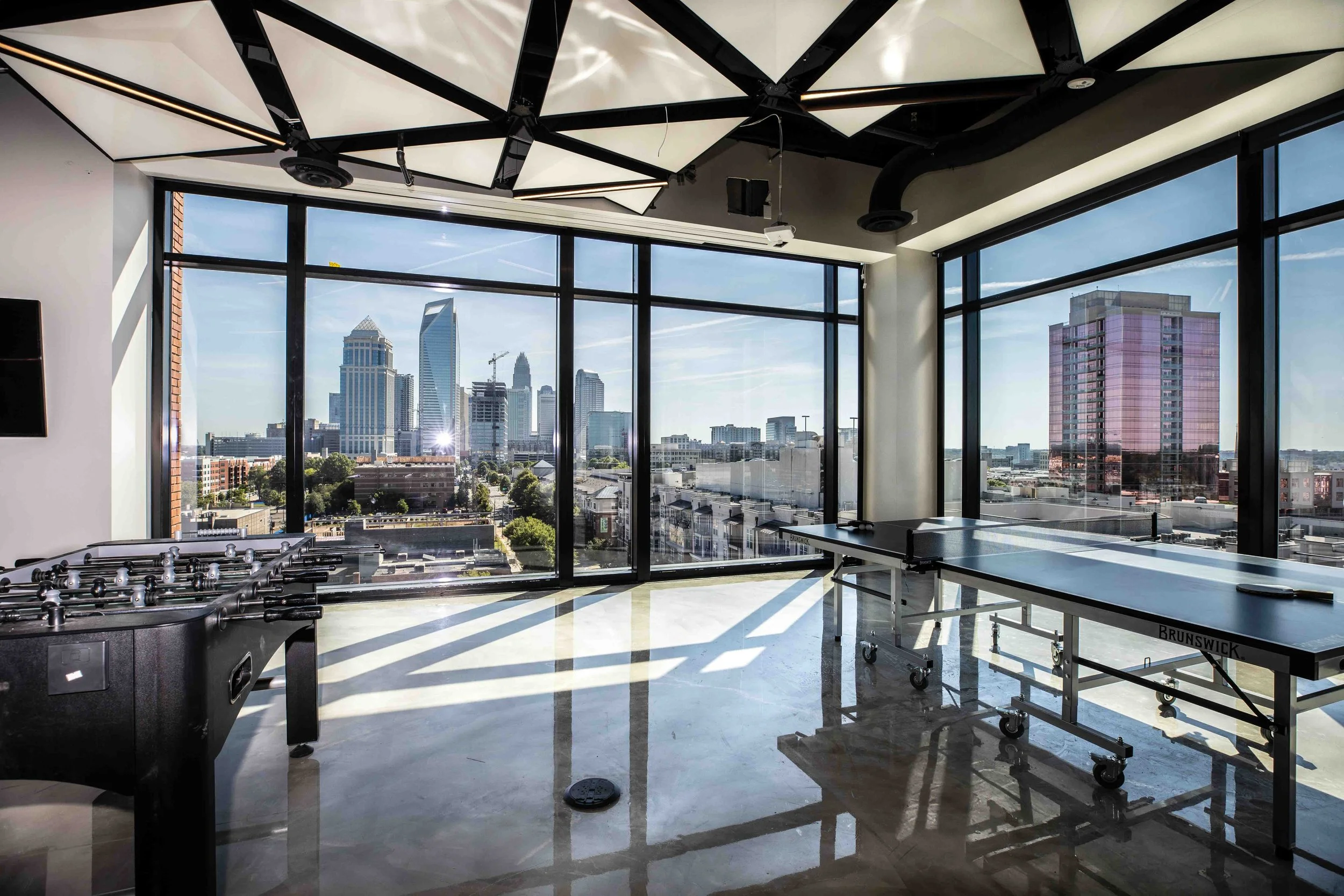 Interior of a modern high-rise building with large floor-to-ceiling windows showing a city skyline, including tall skyscrapers and blue sky. The room has a foosball table and a ping pong table, with shadows cast on the marble floor from sunlight.