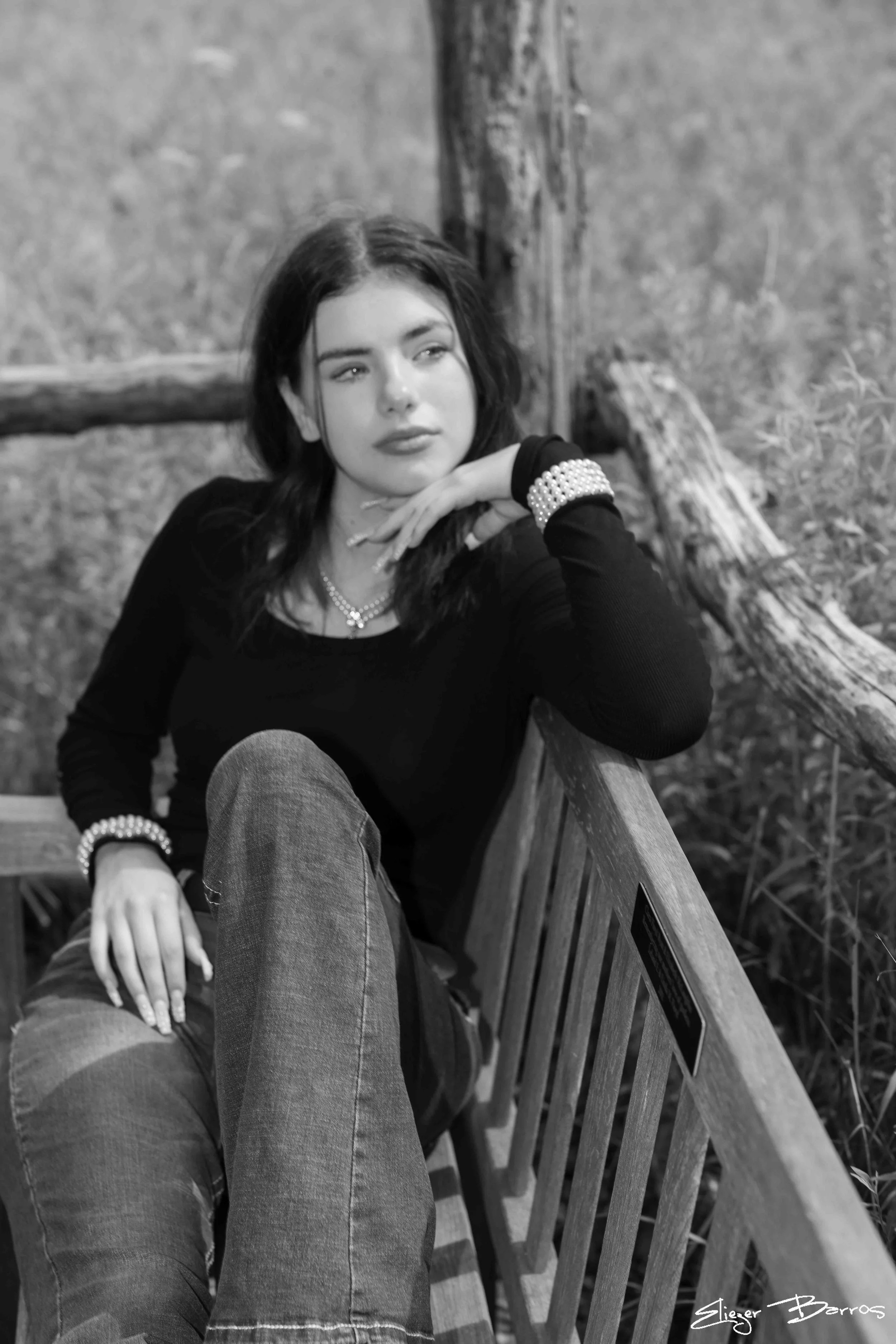 A black and white photo of a young woman with dark hair, wearing a black top, jeans, and pearl jewelry, sitting on a wooden bench outdoors with trees and grass in the background, resting her chin on her hand and gazing to the side.