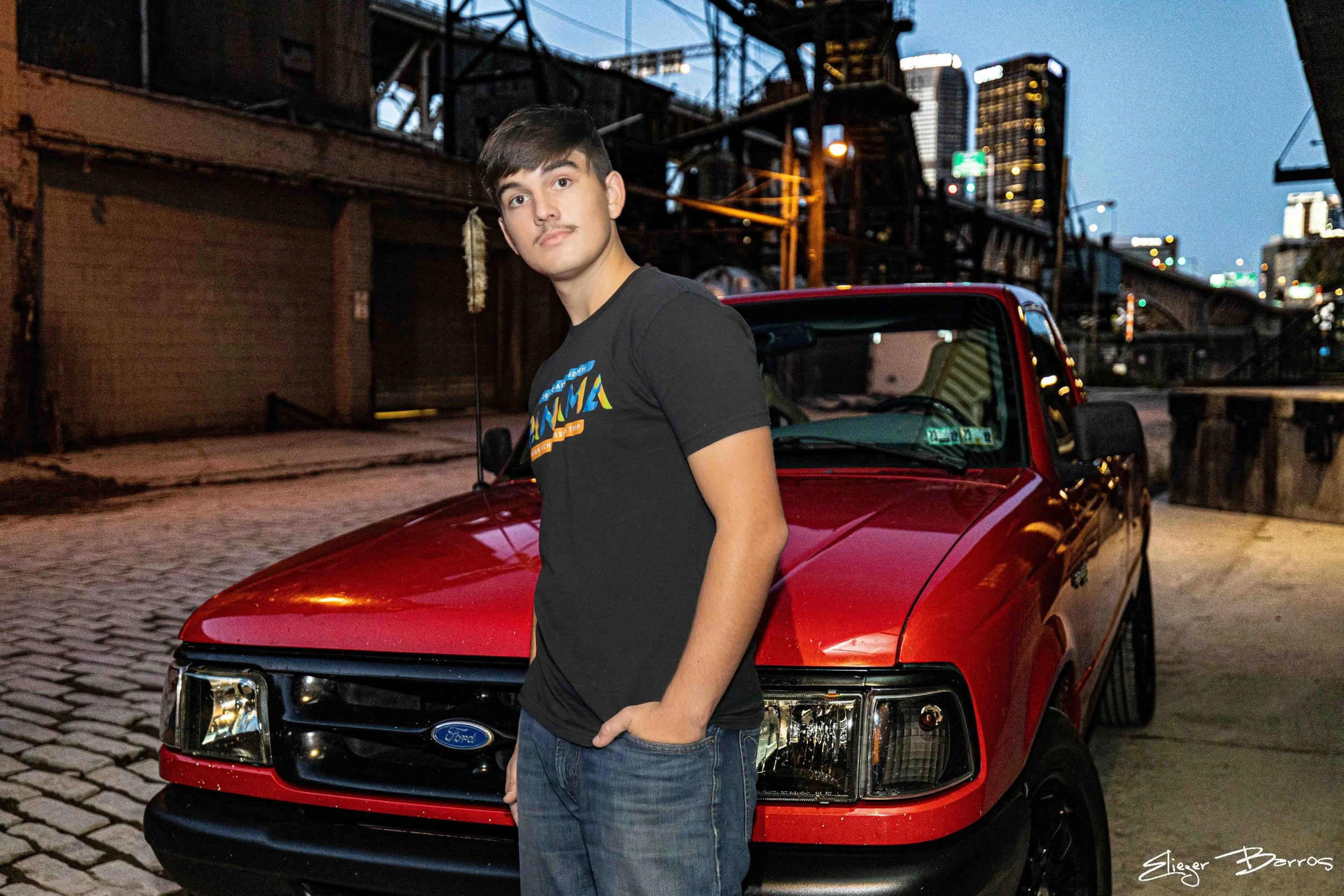 A young man with dark hair and a mustache standing in front of a red Ford pickup truck on a brick-paved street during dusk. Tall city buildings and an industrial bridge structure can be seen in the background.
