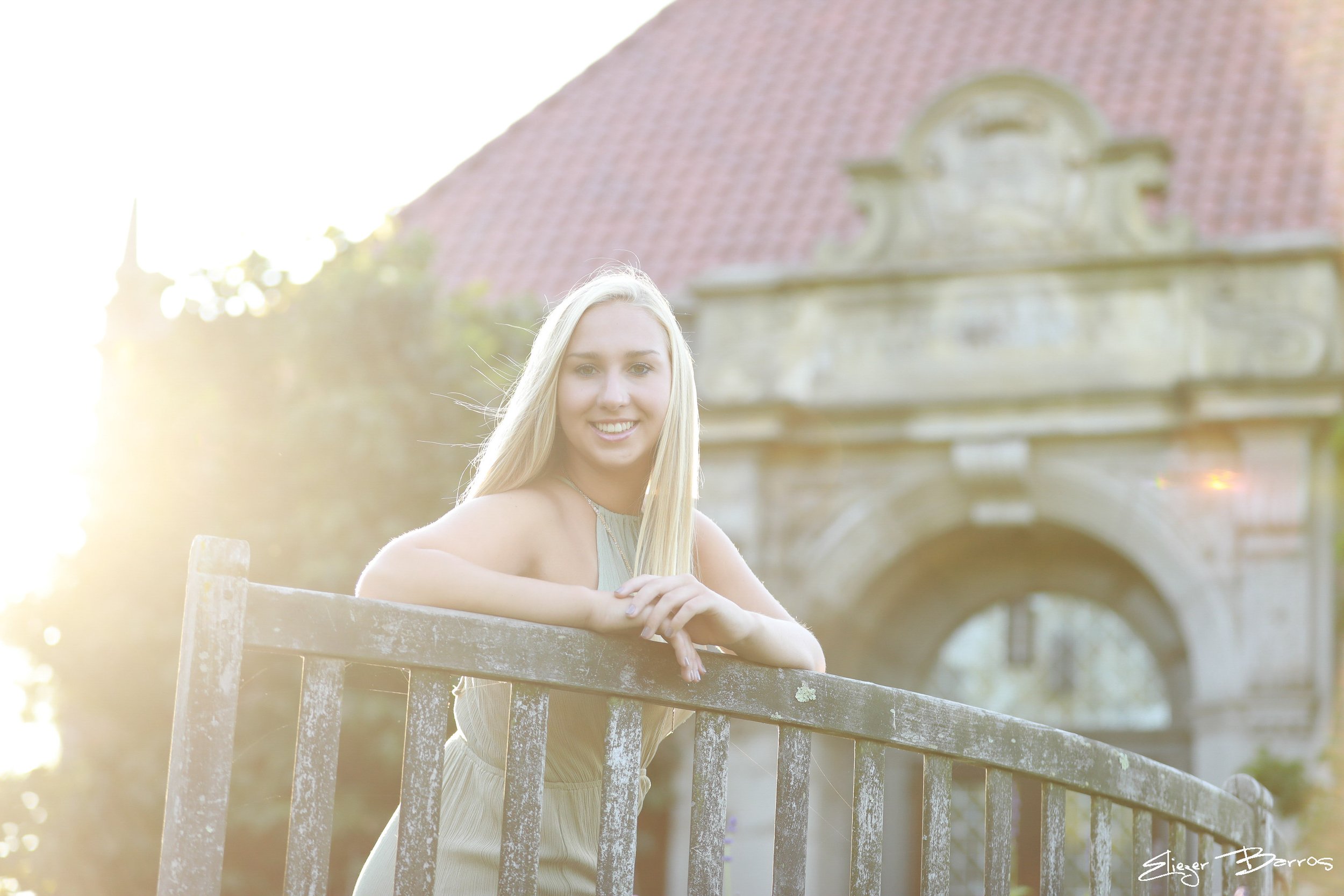 A smiling young blonde woman leaning on a wooden fence outdoors, with a historic stone bridge and roof in the background, illuminated by warm sunlight.