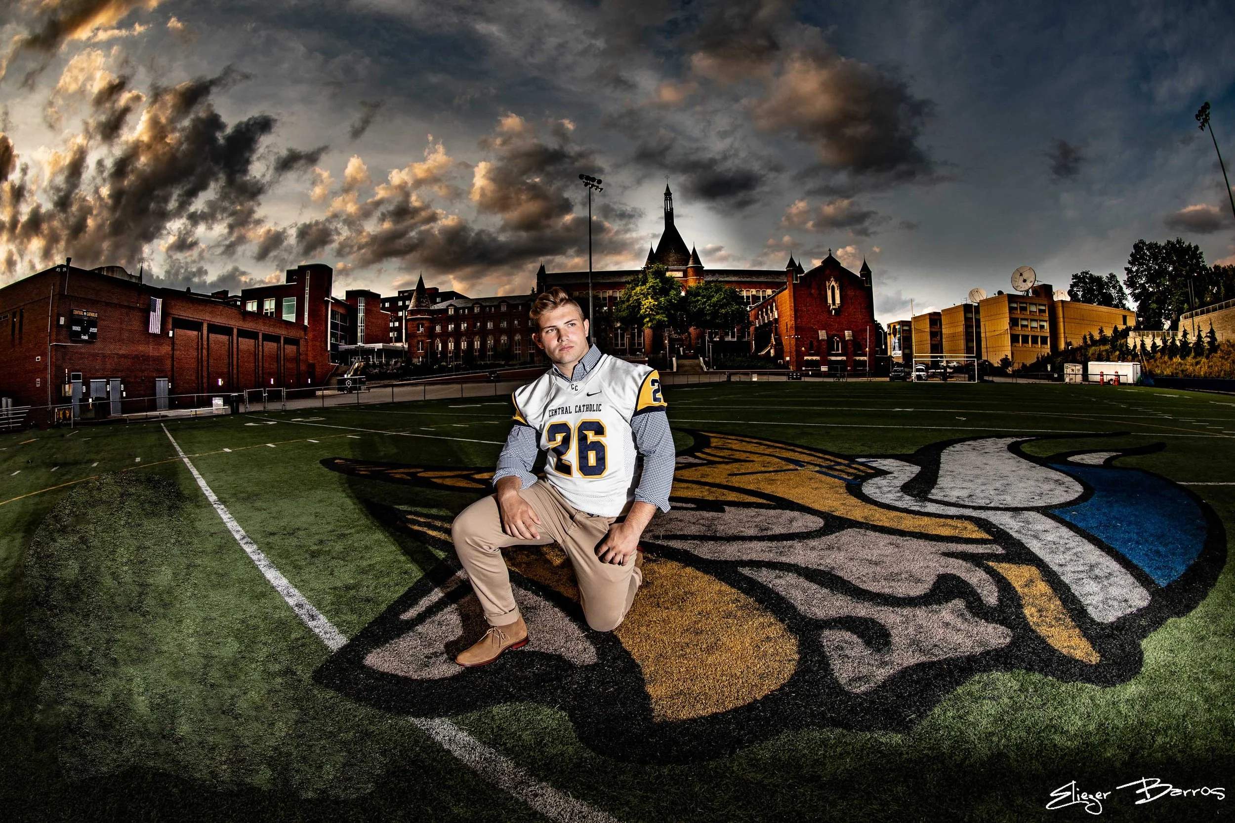 A young man kneeling on a football field with a sports jersey, set against a dramatic cloudy sky and city buildings in the background.