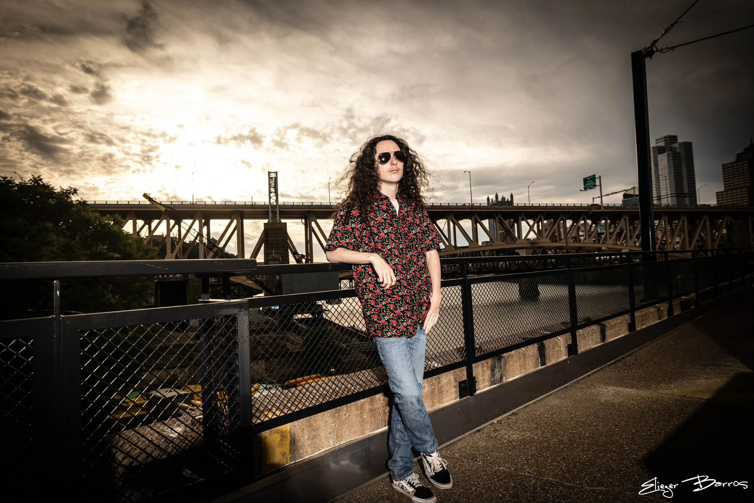 A woman with curly hair wearing sunglasses, a floral shirt, and jeans standing by a metal railing on a bridge, with a city skyline and a bridge in the background under a cloudy sky.