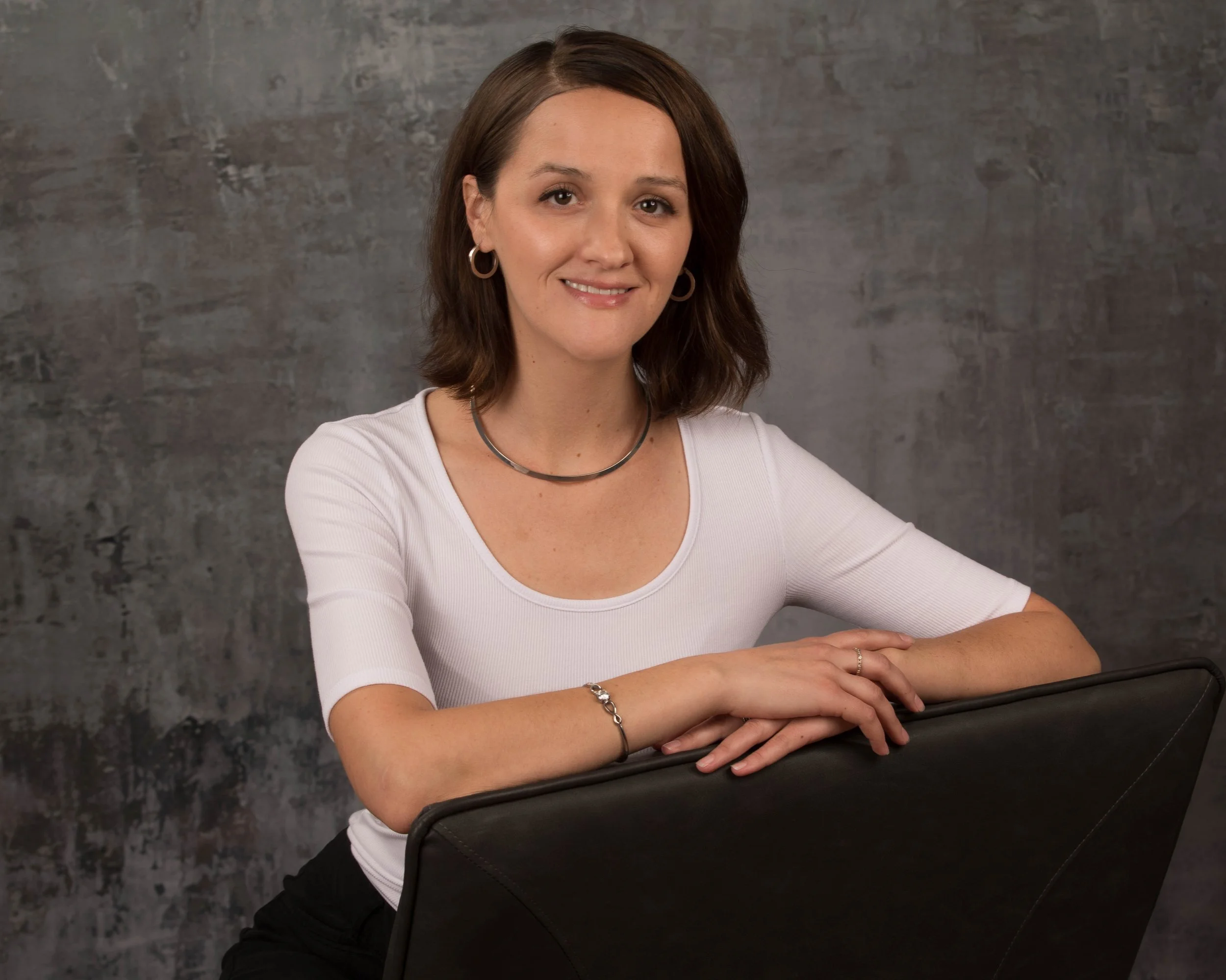 A woman with shoulder-length brown hair, wearing a white top, silver hoop earrings, a silver necklace, and a bracelet, is sitting at a table with her hands resting on it. She is smiling slightly and looking at the camera. The background is a textured