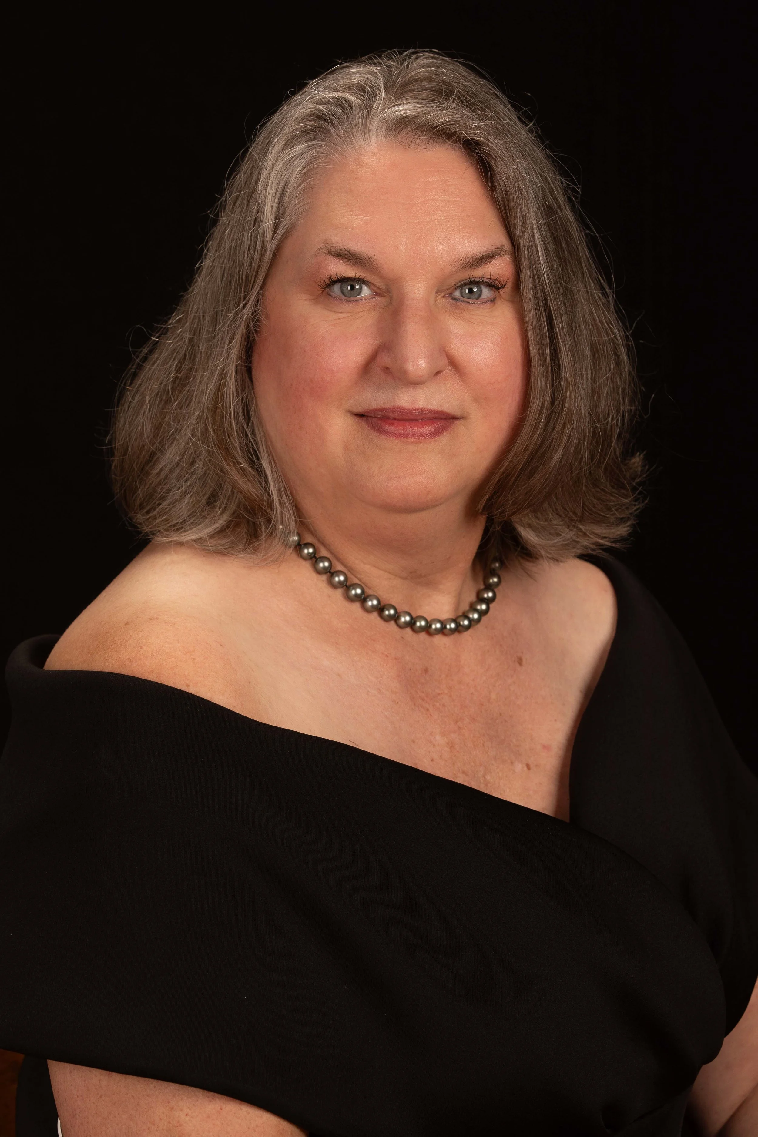 Close-up portrait of a woman with shoulder-length gray hair, blue eyes, and light skin, wearing a black off-shoulder dress and a pearl necklace, against a black background.