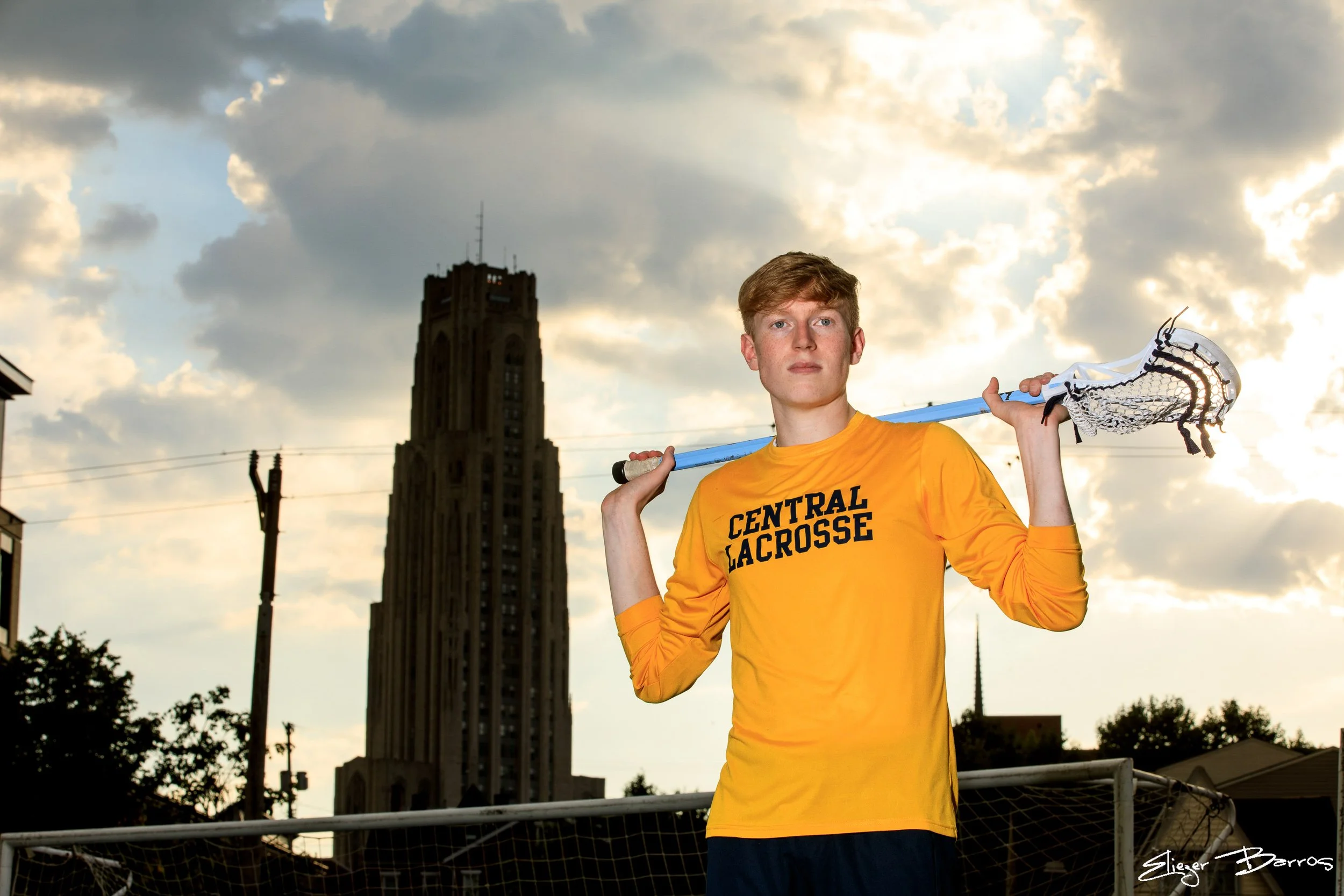Young male lacrosse player in yellow shirt holding lacrosse stick over shoulders, standing outdoors with cloudy sky and a tall building in the background.