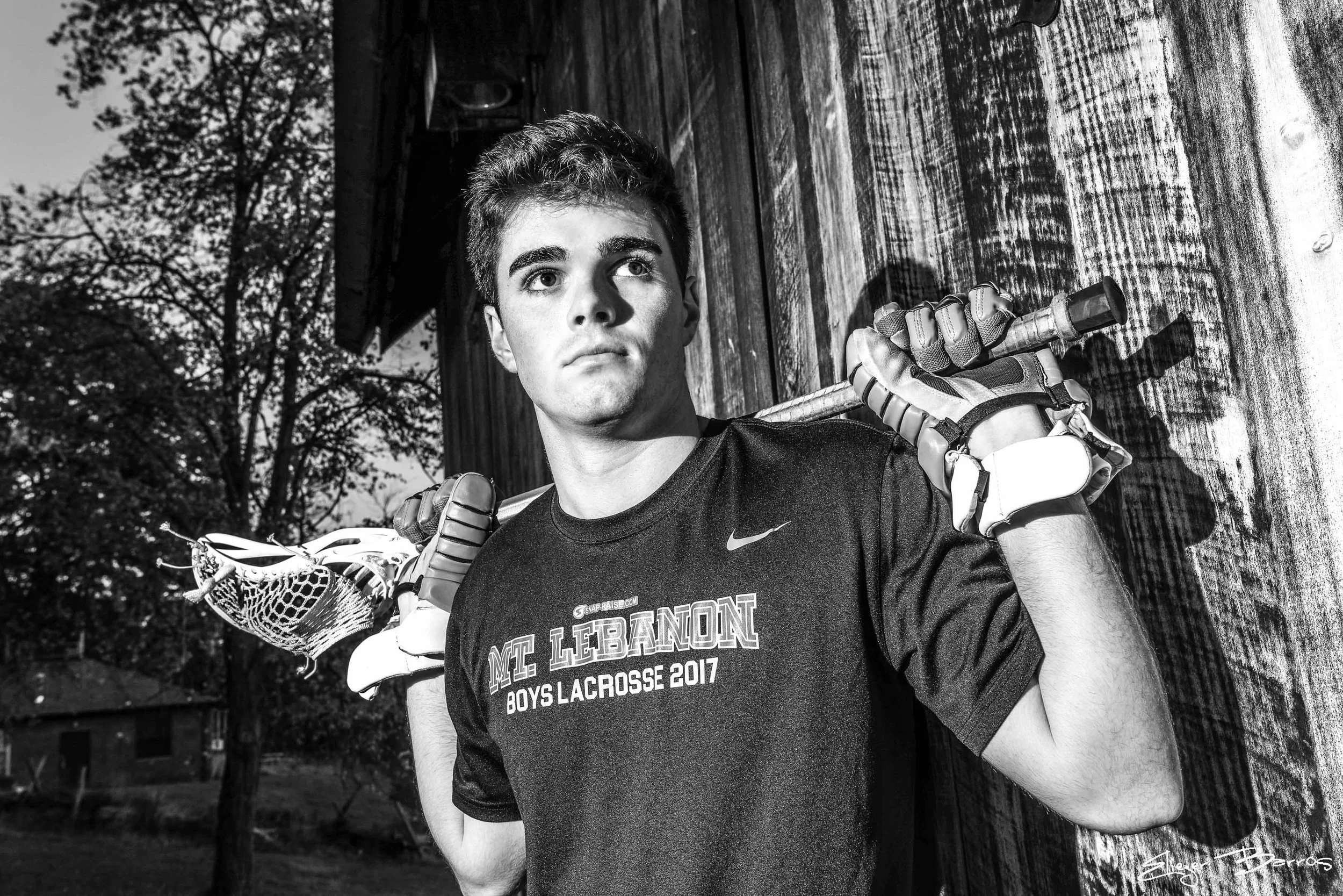 A young man wearing a black t-shirt with text about a lacrosse event, standing outdoors on a wooden surface, holding a lacrosse stick, with trees in the background.