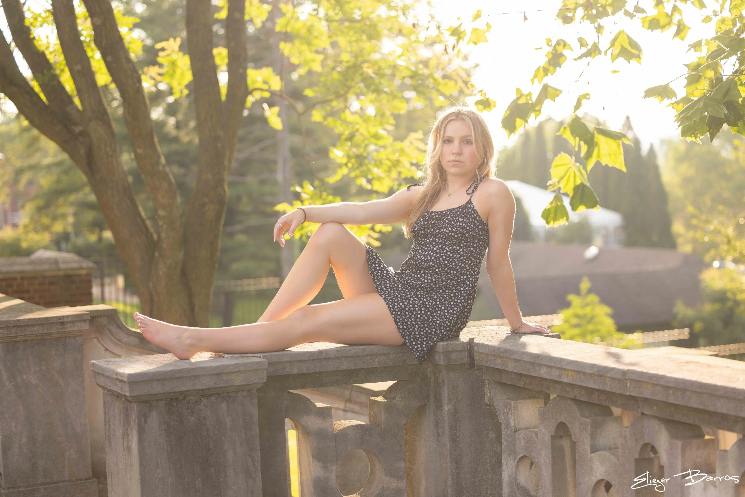 Young woman in a black floral dress sitting on a stone ledge outdoors, with sunlight filtering through green trees in the background.