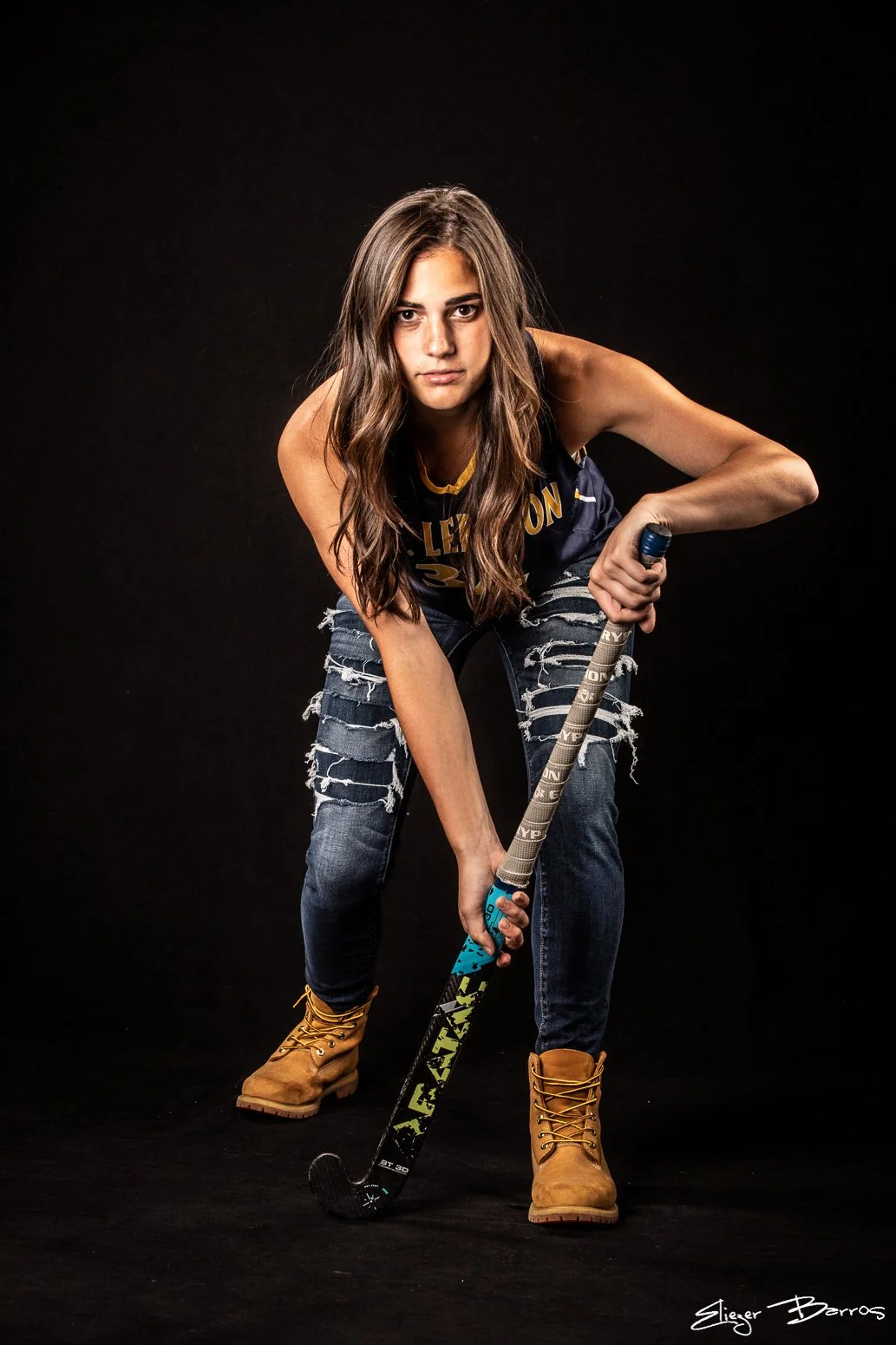 Young woman with long brown hair wearing ripped jeans, a black sports jersey, yellow work boots, and holding a hockey stick in a ready stance against a black background.