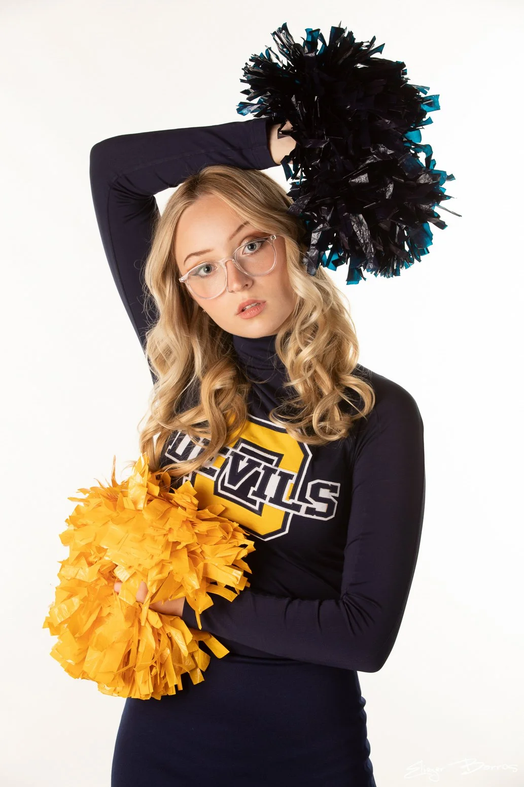 A young woman with glasses and blonde curly hair poses with black and yellow pom-poms, wearing a cheerleading uniform with a yellow and black logo. She stands against a plain white background.
