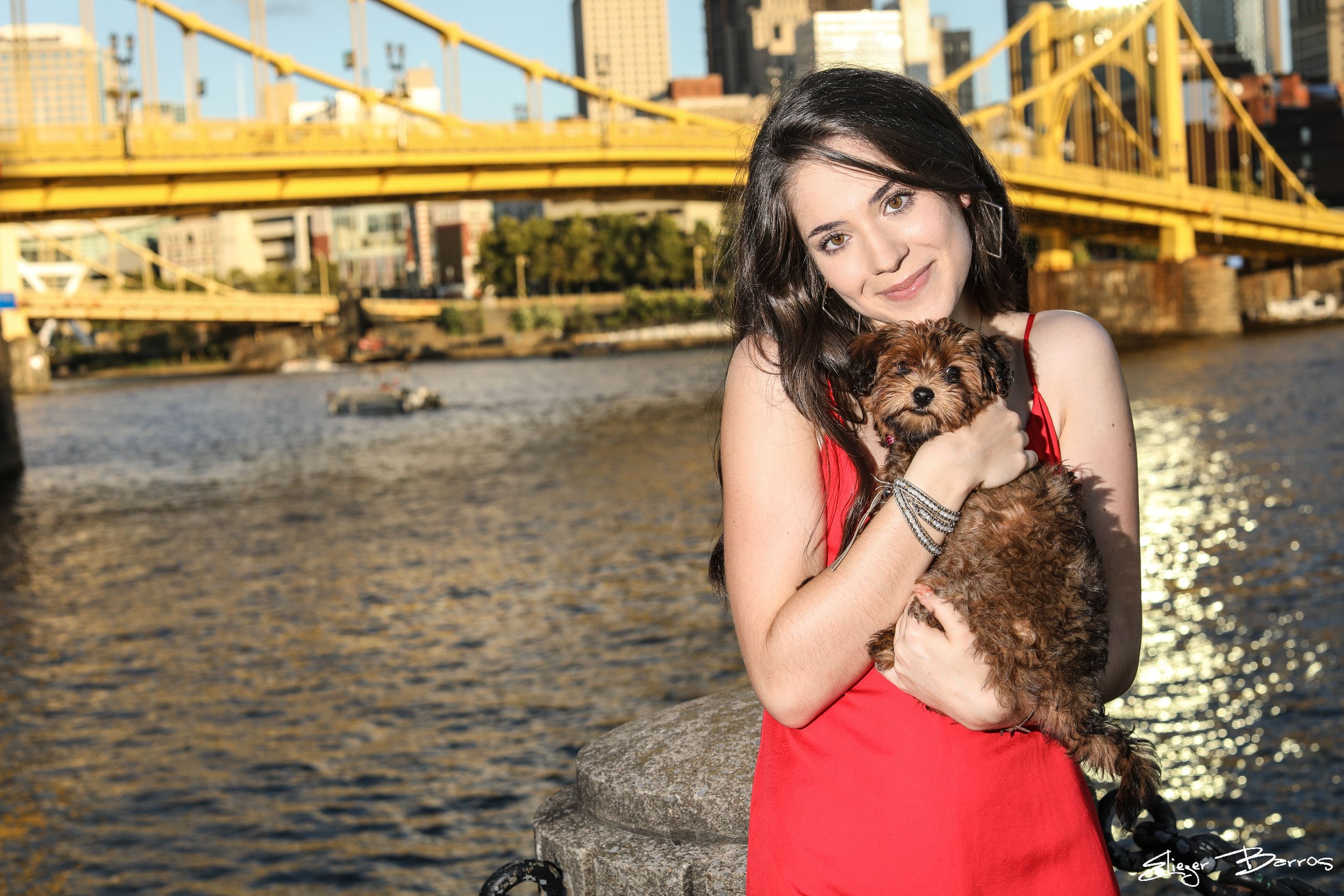 A young woman with dark hair in a red dress holding a small brown puppy by a riverside with a yellow bridge and tall city buildings in the background.