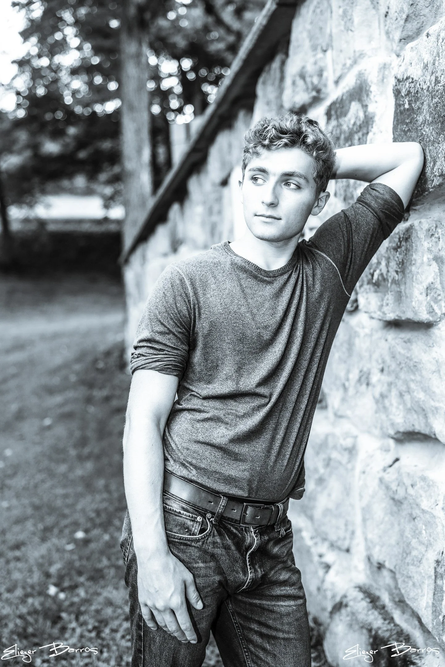 Black and white photo of a young man leaning against a stone wall outdoors. He is wearing a casual t-shirt and jeans, with one arm resting behind his head and the other hanging down. There are trees and a blurry background.