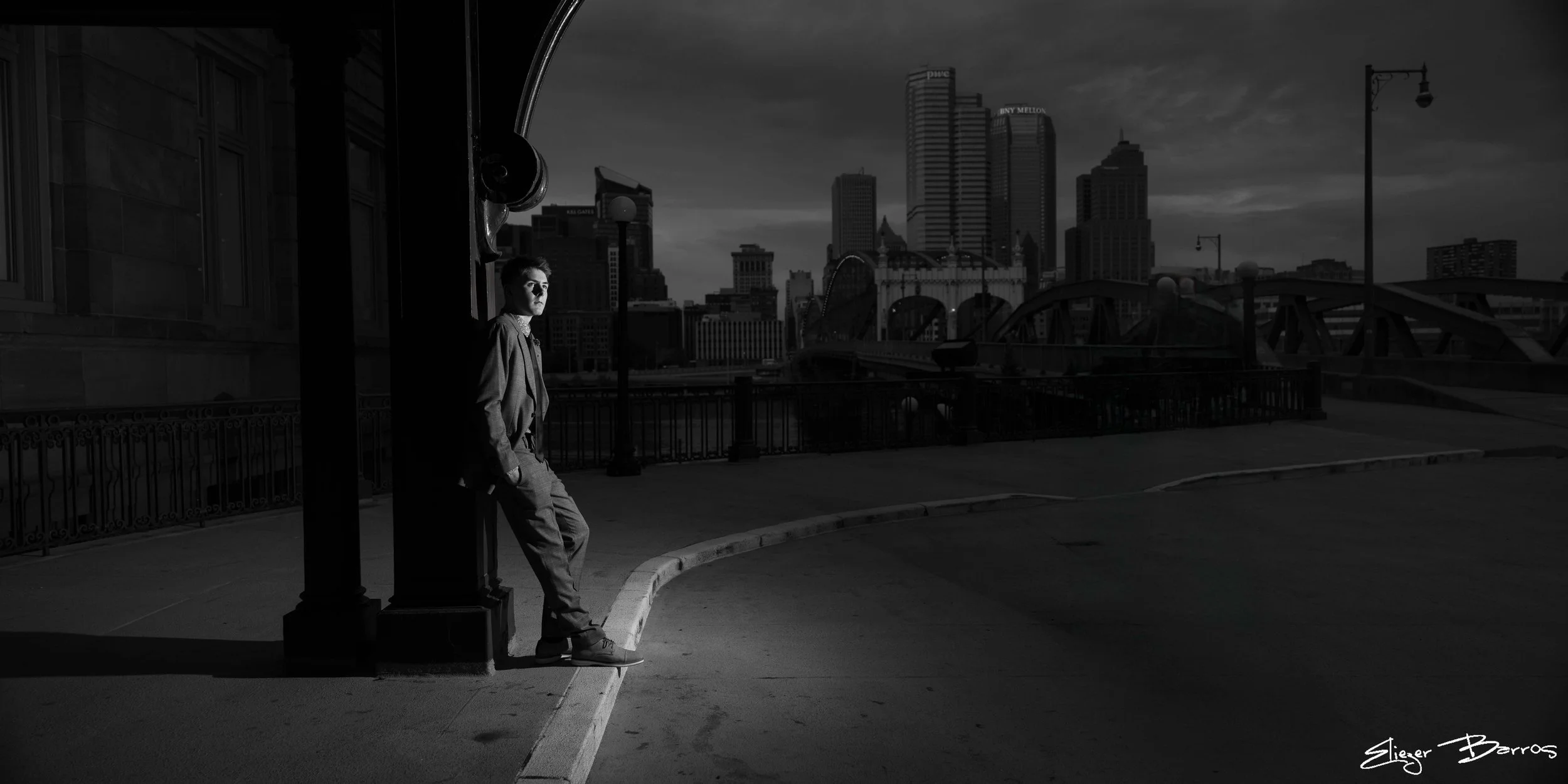 A man in a suit leaning against a lamp post on a city bridge at dusk, with a city skyline and tall buildings in the background, in black and white.