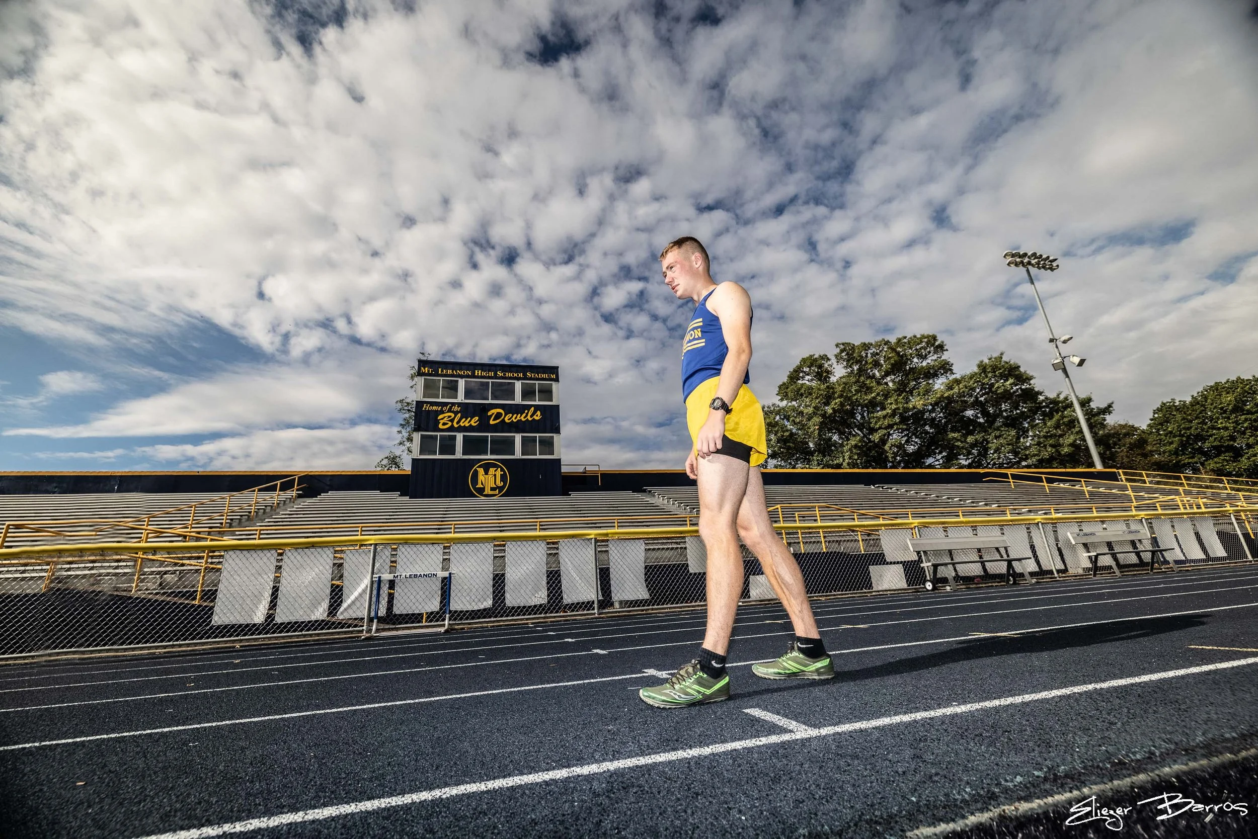 A male athlete in yellow shorts and a blue tank top stands on a running track at Mt. Lebanon High School Stadium. The stadium has empty bleachers, a scoreboard, and a large sign that reads "Home of the Blue Devils." The sky above is partly cloudy wit