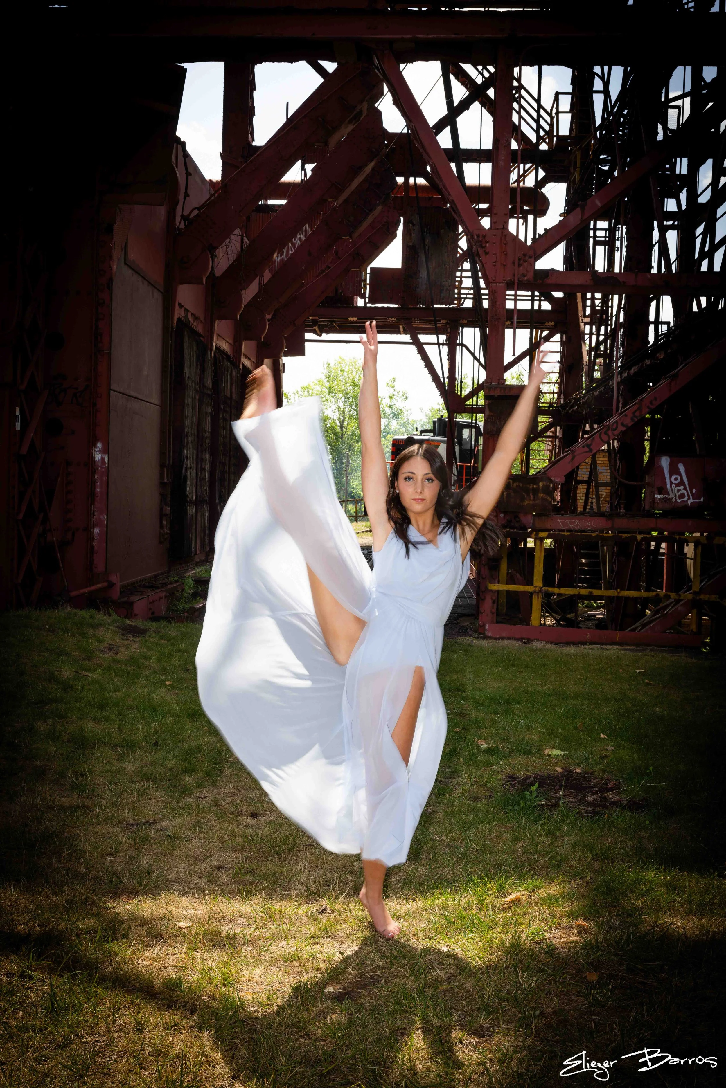 A woman in a white dress performing a dance move with one leg raised and her arms extended upwards, standing outdoors in front of an old, rusty industrial structure with metal stairs and beams, under a partly cloudy sky.