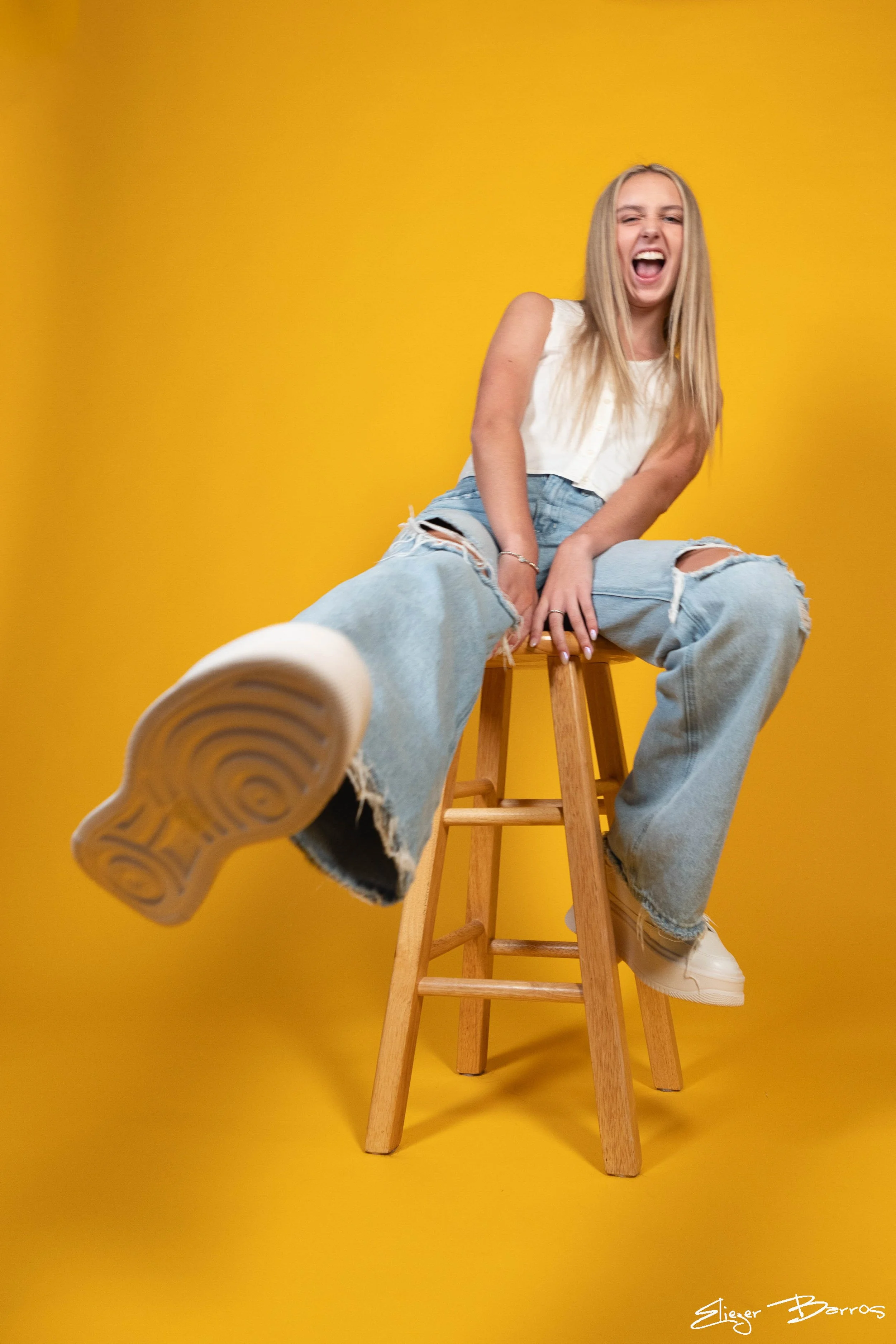 A young woman with long blond hair, wearing a white sleeveless top and ripped jeans, laughing and sitting on a wooden stool against a yellow background. She is leaning back with her legs extended, wearing white sneakers, and has a playful, joyful exp
